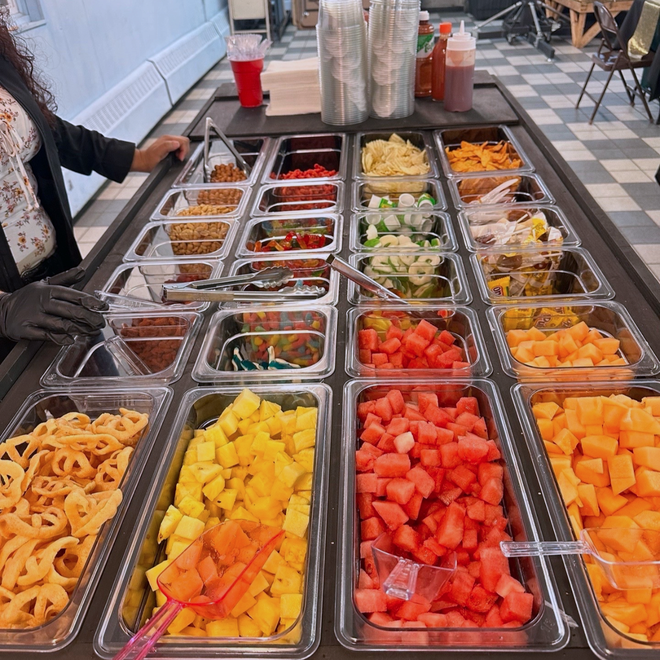 A variety of chopped fresh fruits including watermelon, pineapple, cantaloupe, and honeydew melon in a buffet-style setup. Tongs are visible for serving.