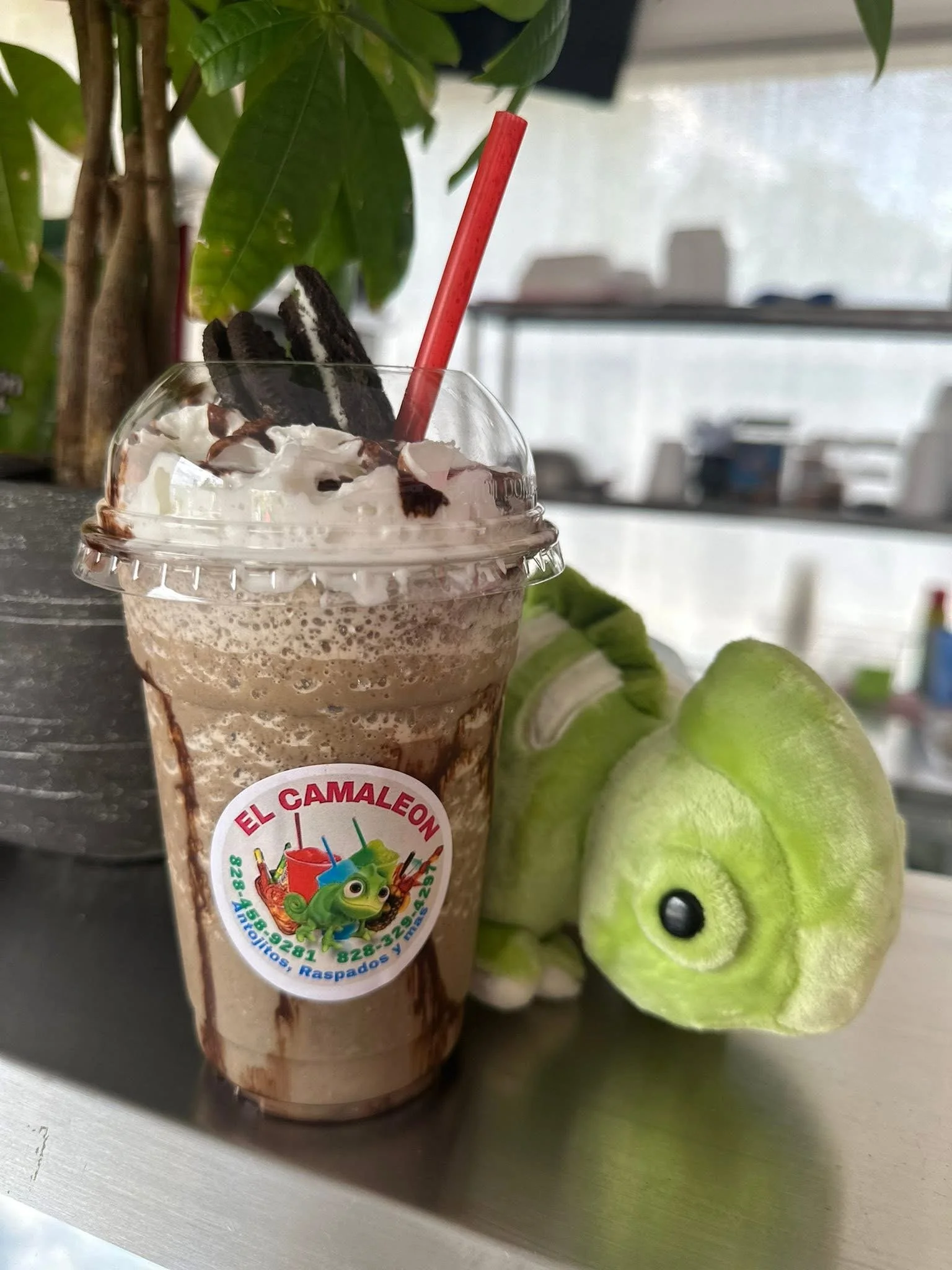 Chocolate milkshake topped with whipped cream, Oreo cookies, and chocolate syrup, with a red straw, placed beside a green plush toy on a metal surface.