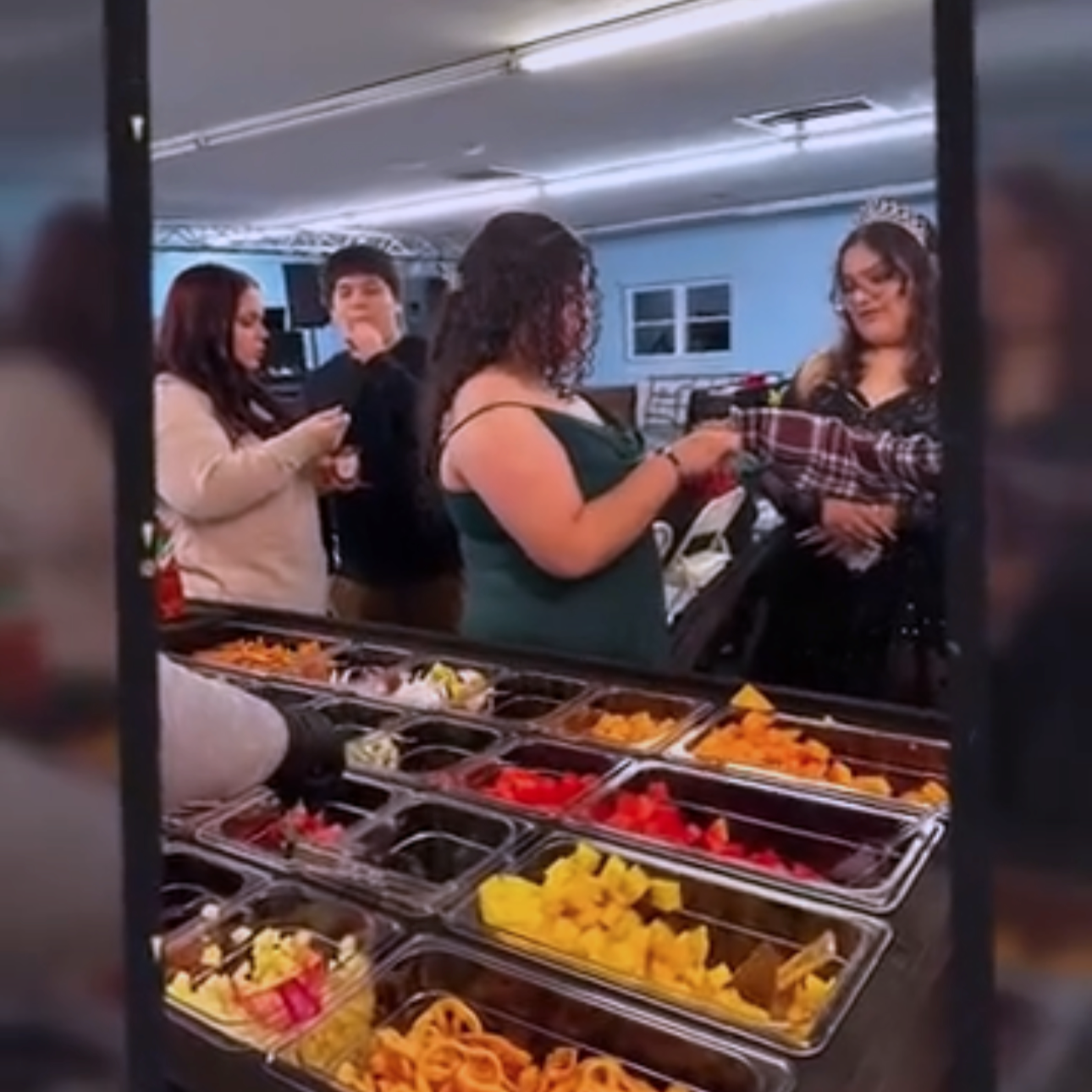Group of young women at a birthday party, one wearing a tiara, standing near a food buffet with various fruit and snack options, set in a decorated indoor space.