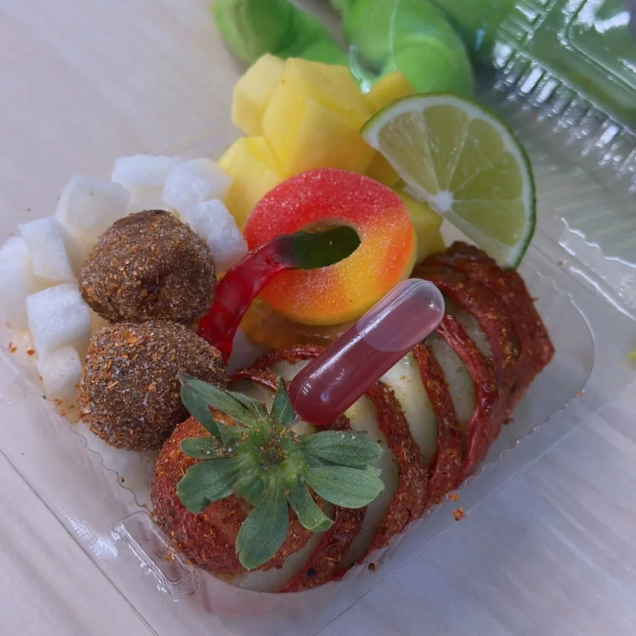 Assorted appetizers including candy coated grapes, diced fruit, a strawberry topped with a chamoy and tajin, a lime wedge, and a red spiral-cut cucumber snack wrapped in tamarind candy, in a clear plastic container.