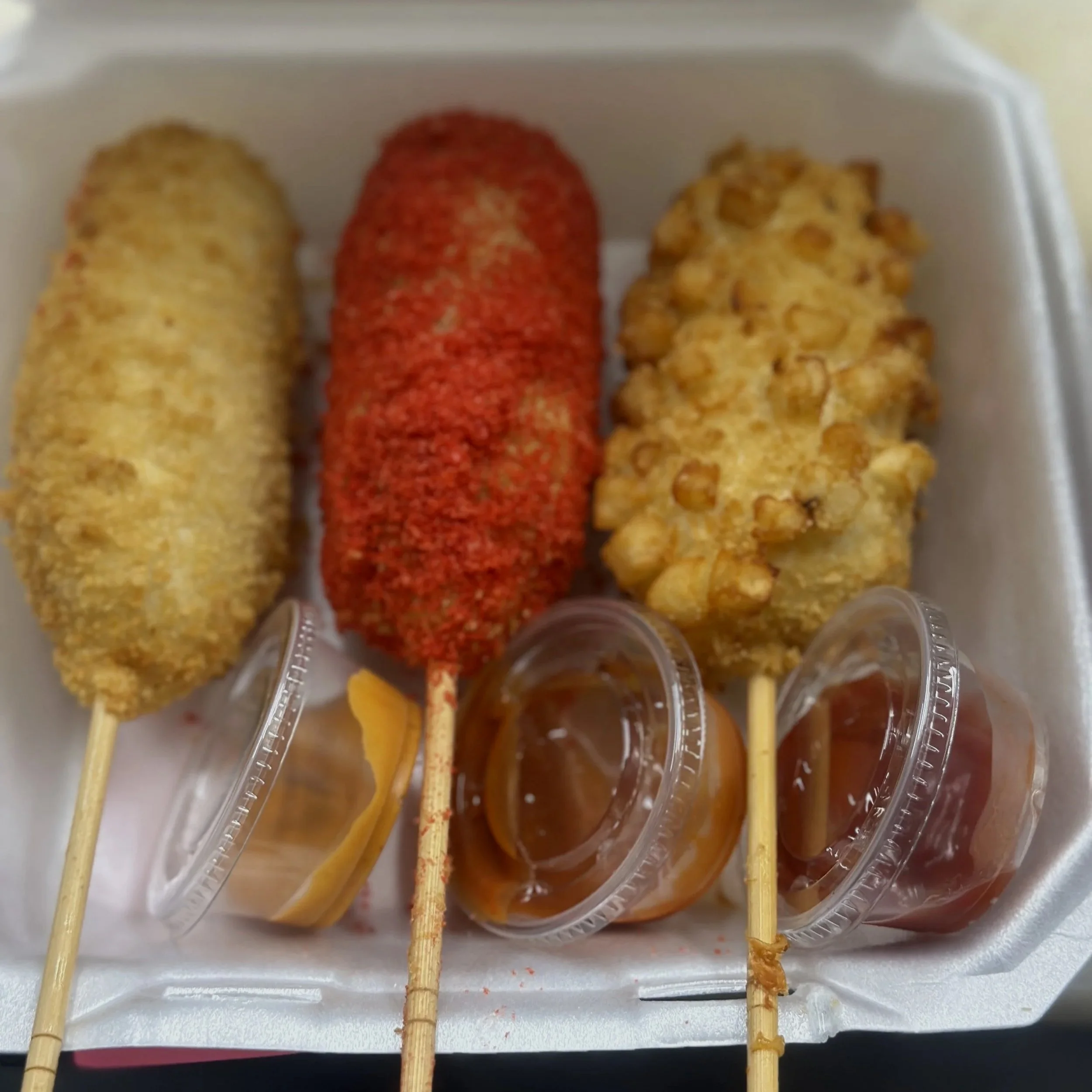 Three corn dogs with different coatings and dipping sauce in a white foam container.