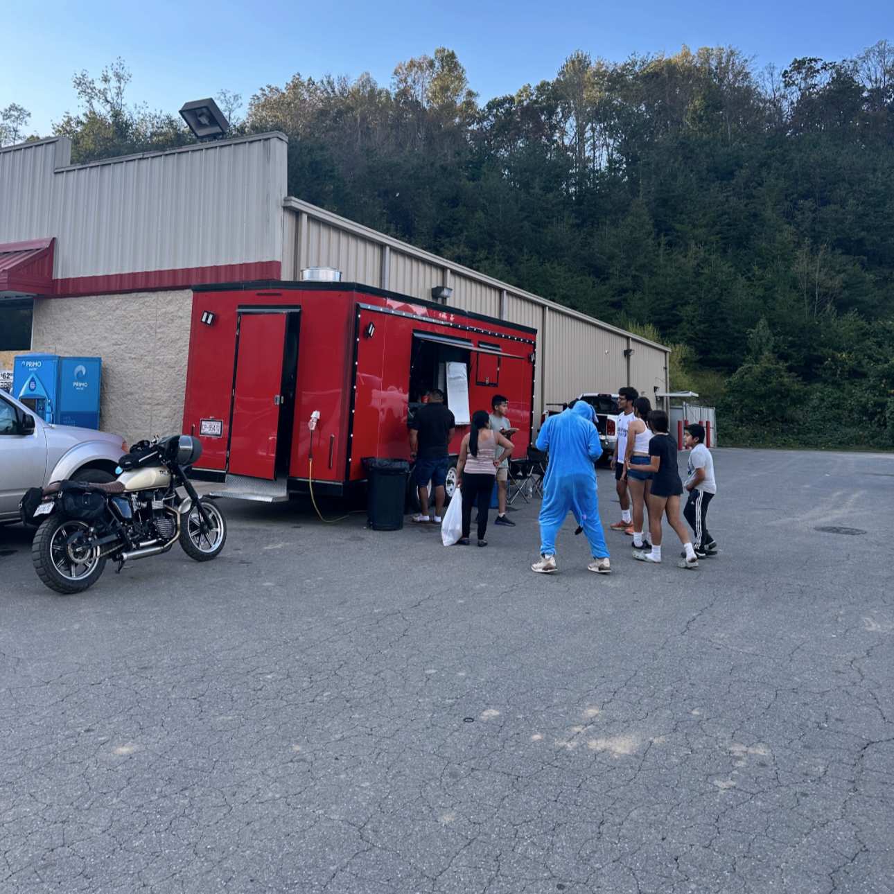 Group of people gathered outside a red food truck with a window, in a parking lot with a motorcycle and SUV, against a background of trees and a blue sky.