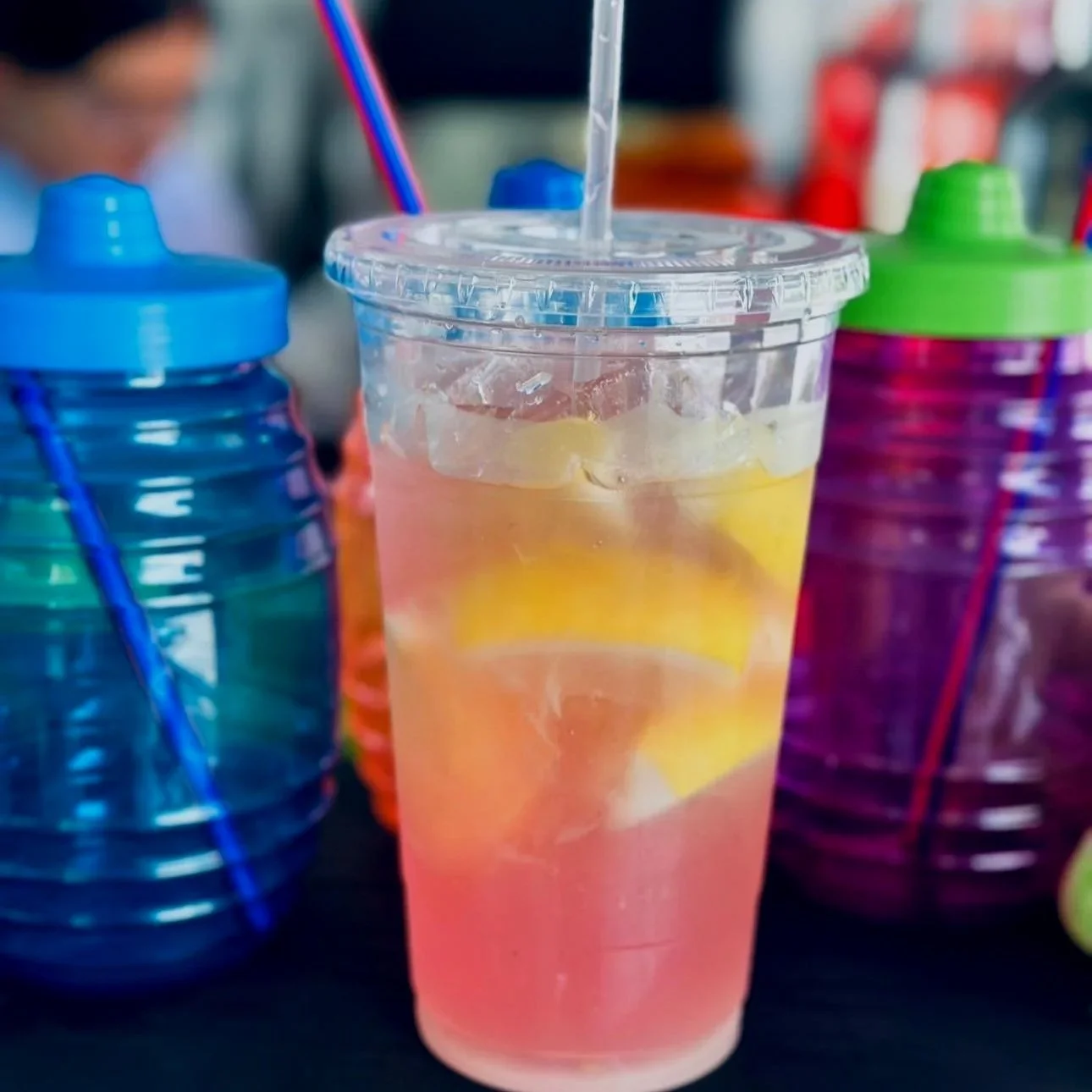 A clear plastic cup with a lid and straw contains pink lemonade with ice and lemon slices, positioned on a table with colorful bottles in the background.