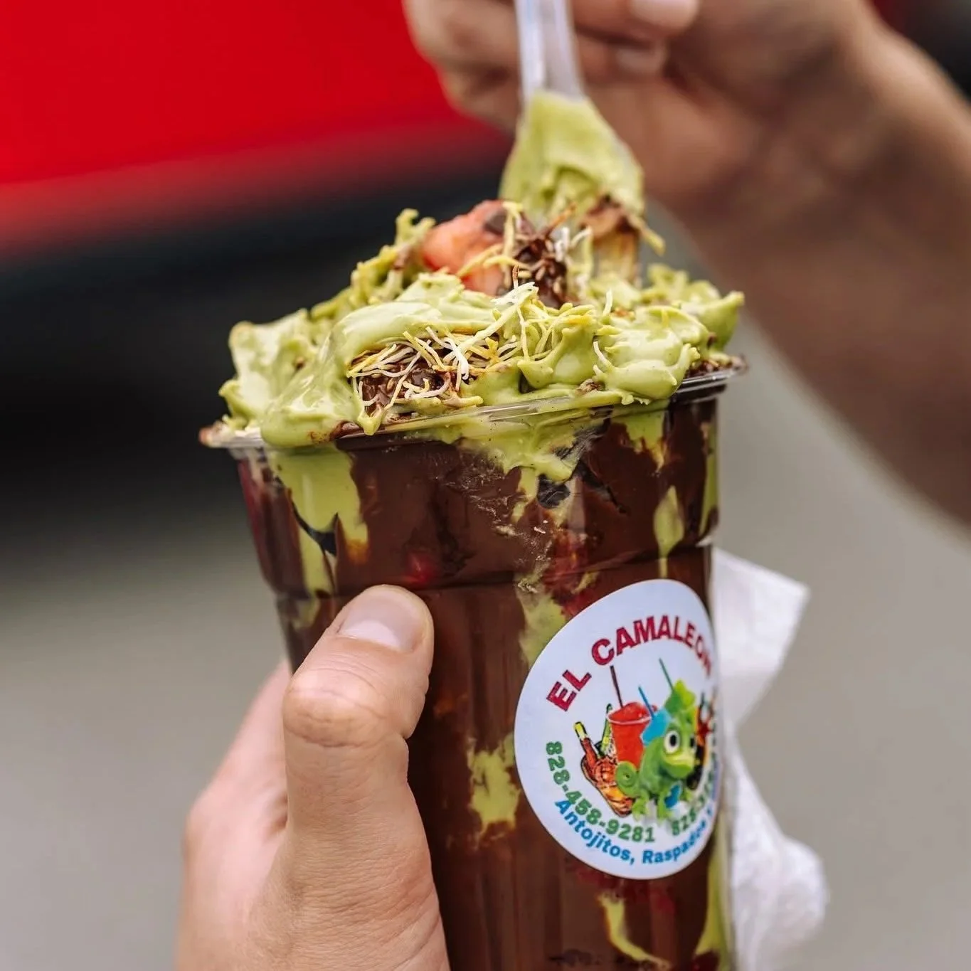A hand holding a Mexican ice cream cup with chocolate and green sauce, topped with avocado, sprouts, and seafood, from El Amale's, displaying a colorful logo with a cartoon character.