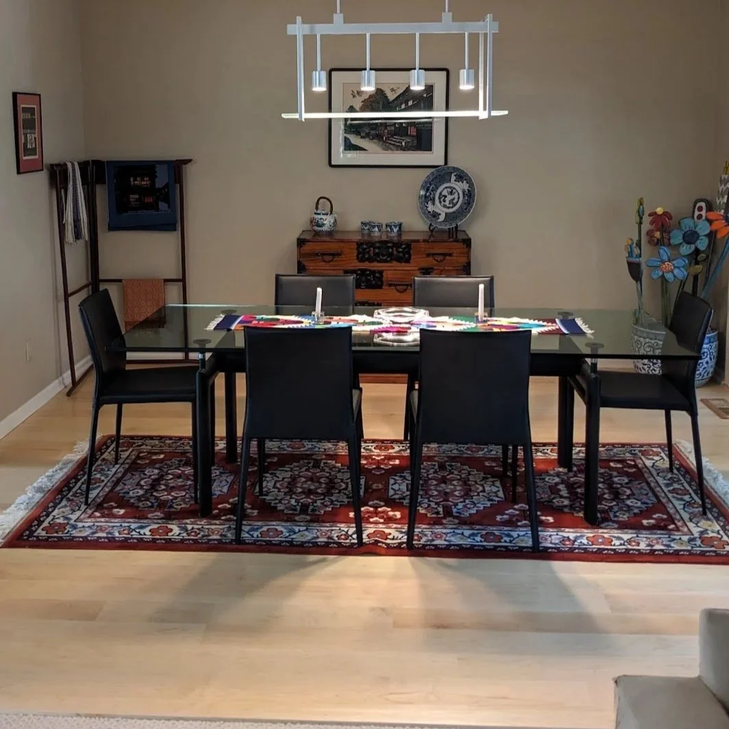 Dining room with a glass table, six black chairs, a red patterned area rug, a wooden sideboard, decorative plates, framed artwork, and a large colorful flower arrangement in a vase.