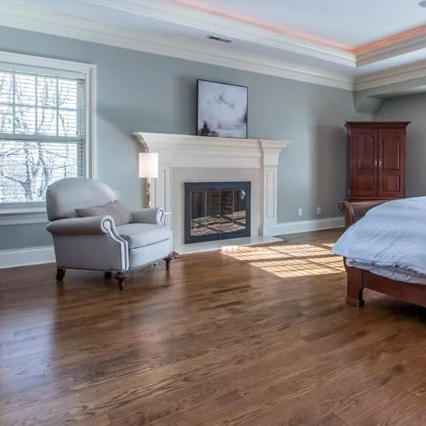 A spacious bedroom with hardwood floors, a white armchair near a window, a fireplace with a white mantel, a bed with white bedding, a wooden wardrobe, and a painting above the fireplace.