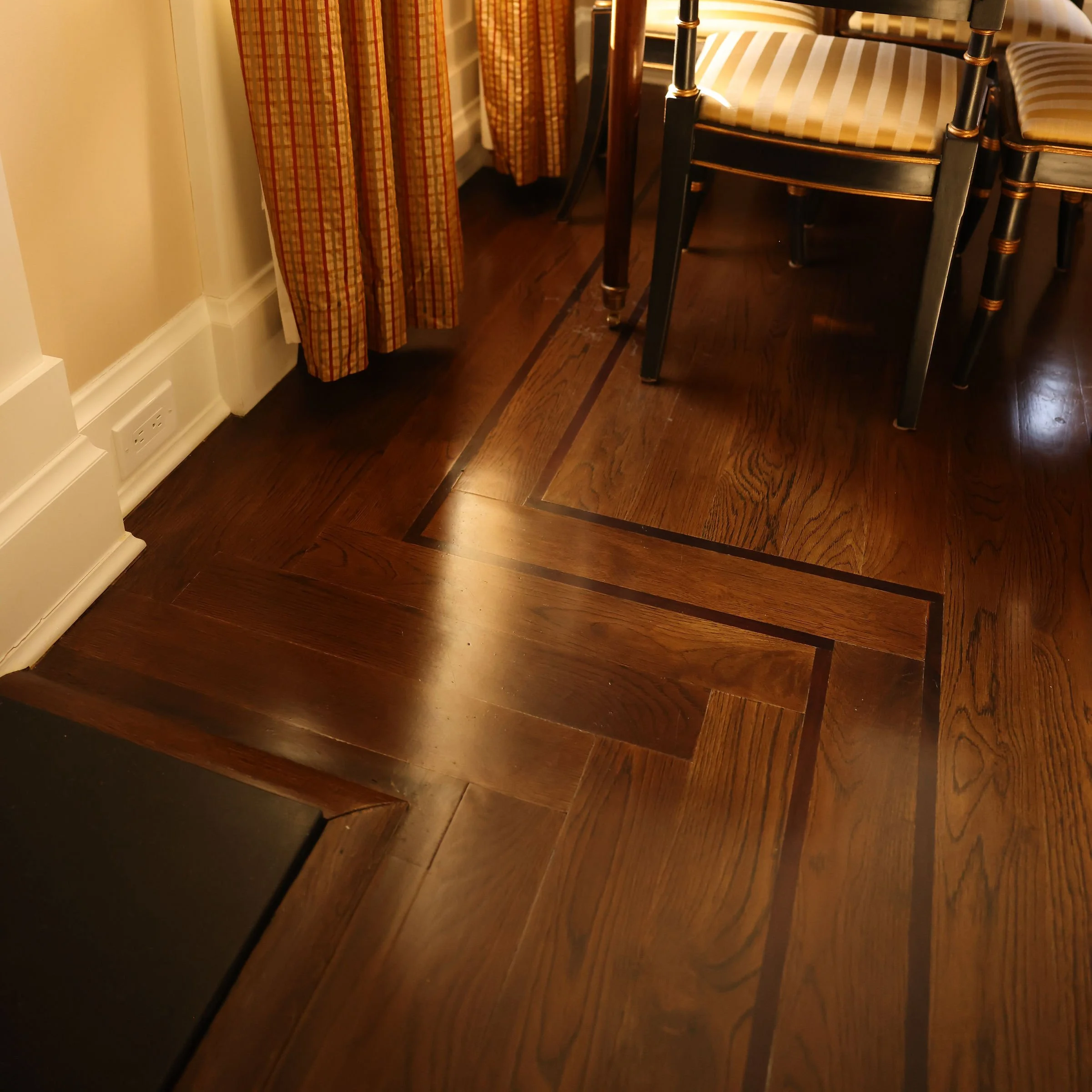 Close-up of a hardwood floor with a decorative border, part of a dining room with chairs and curtains.