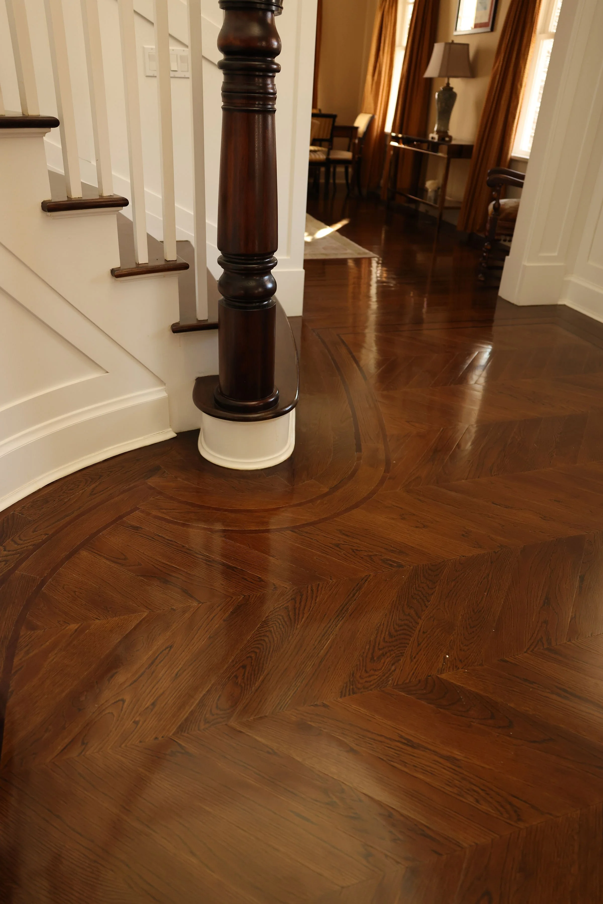 Close-up of a polished hardwood floor with intricate inlaid wood patterns, a wooden staircase post, and part of a curved wall.