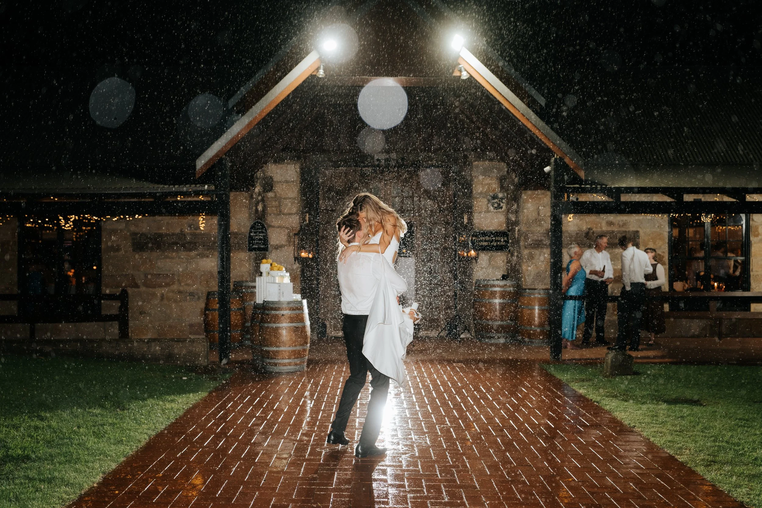 A couple dancing in the rain at night outside a rustic venue, with people mingling in the background.