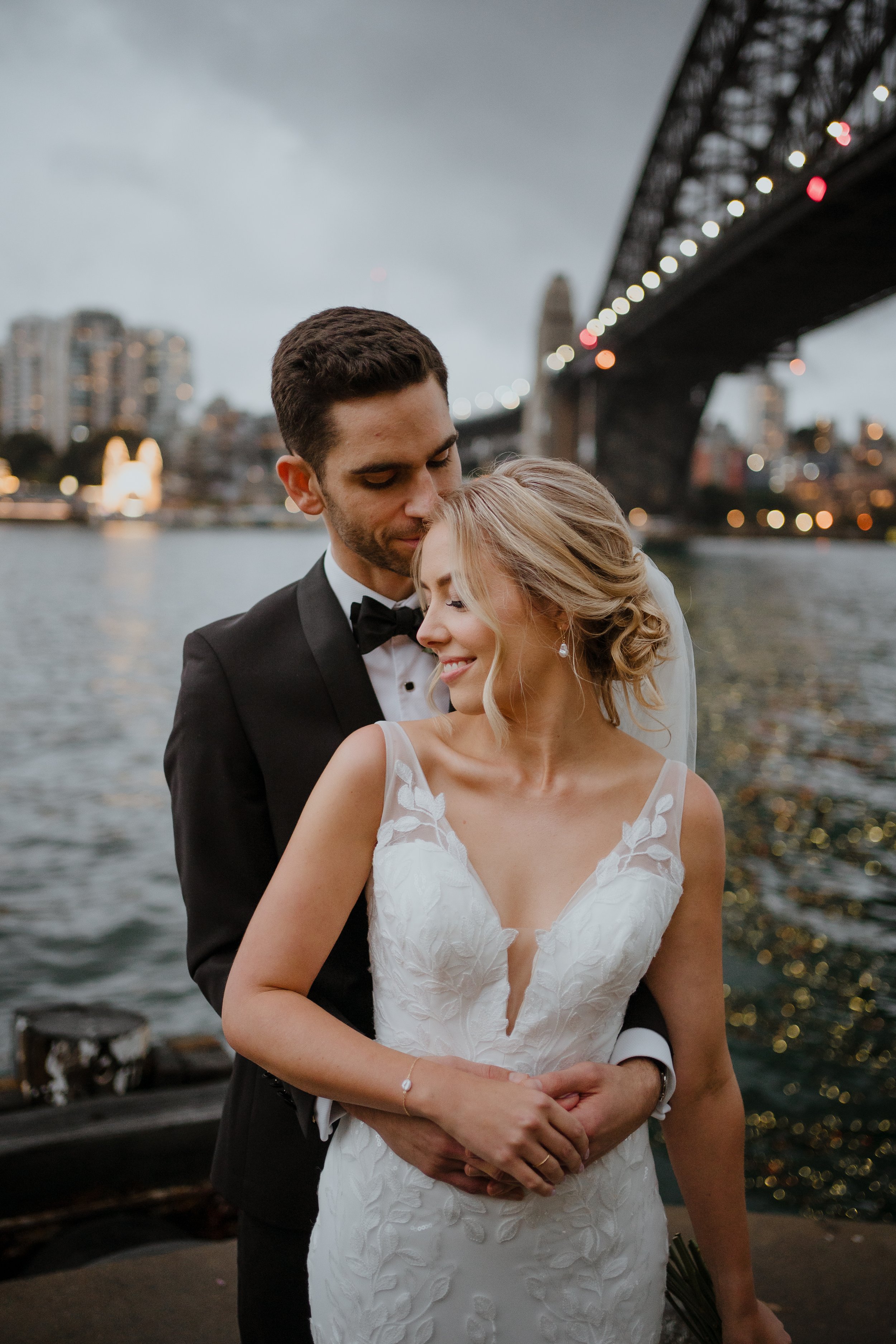 A newlywed couple dressed in wedding attire standing by a river, with a city bridge and skyline in the background during evening.