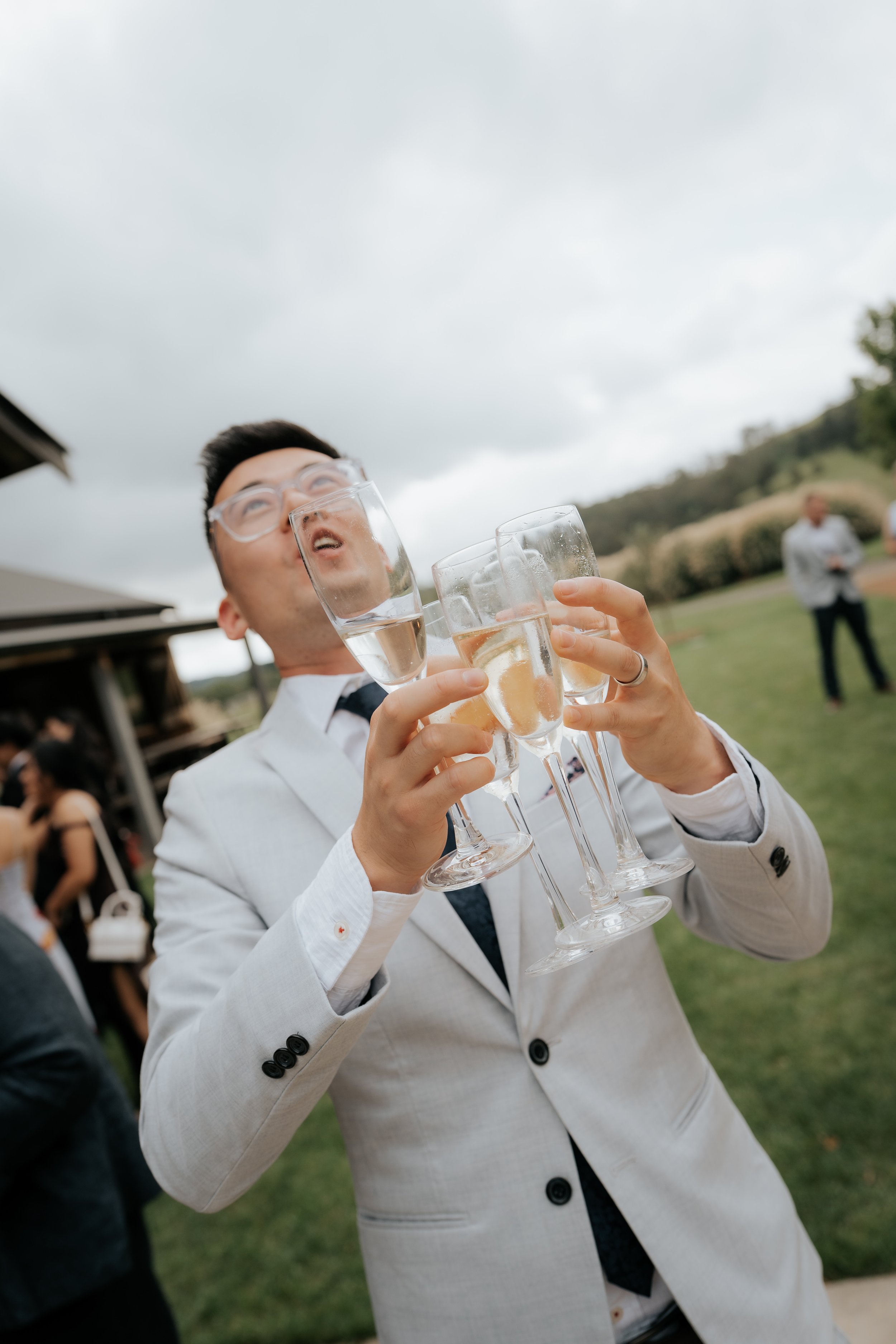 Man in formal attire holding multiple glasses of champagne at an outdoor event.