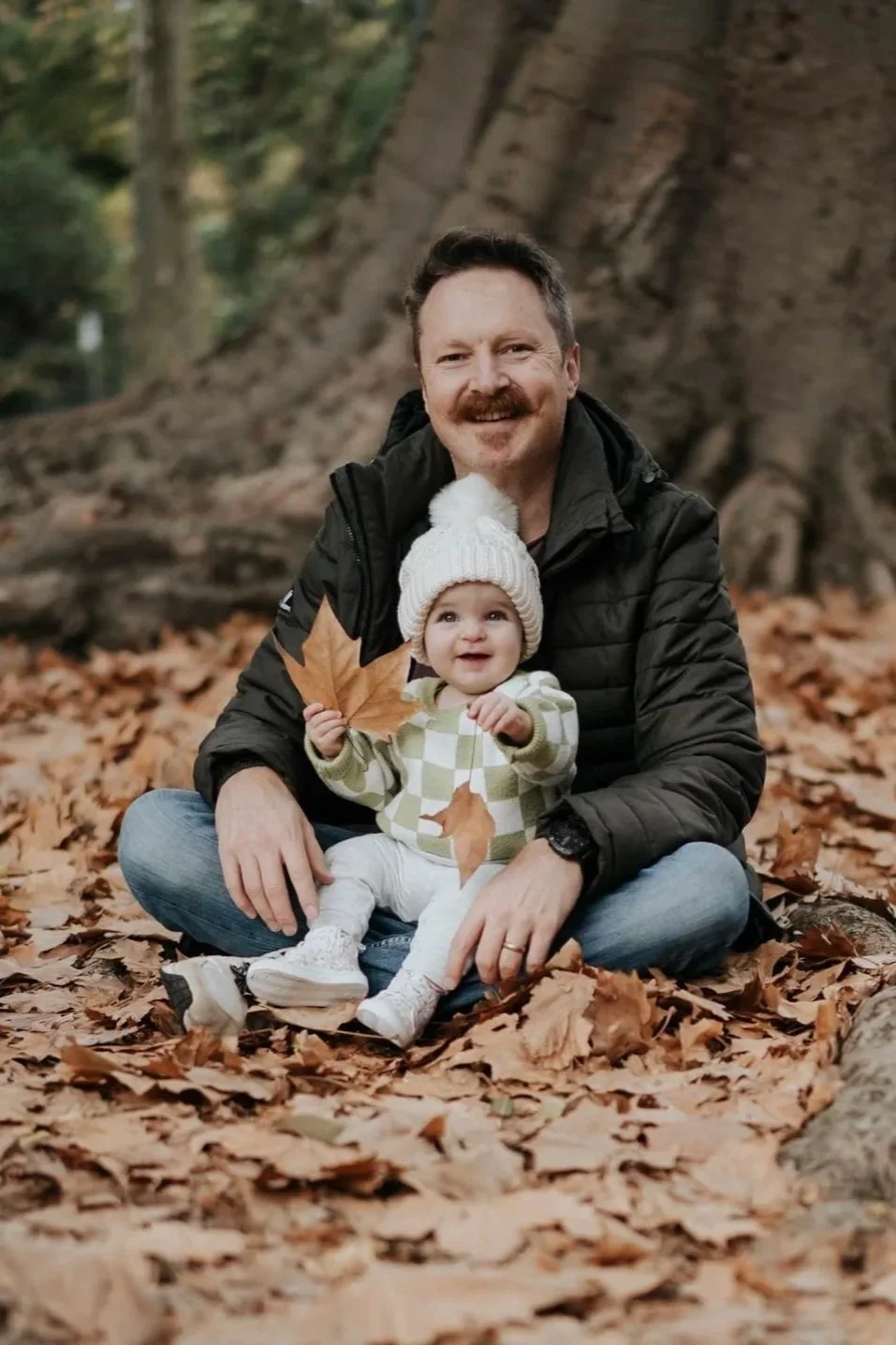 A man and a young girl sitting on fallen autumn leaves in a forest, with the man holding the girl who is holding a large autumn leaf, both smiling.