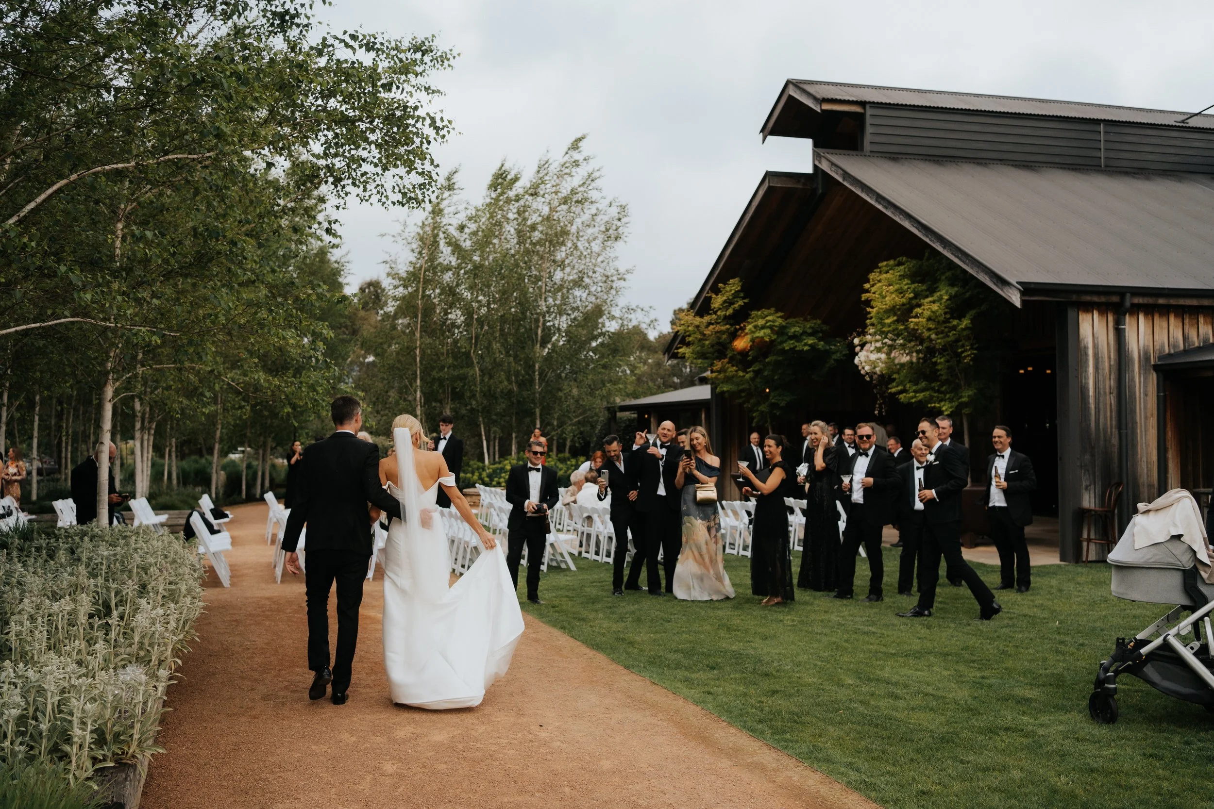 A bride and groom walking down an outdoor aisle surrounded by friends and family in formal attire, with trees and a modern wooden building in the background, during a wedding celebration.