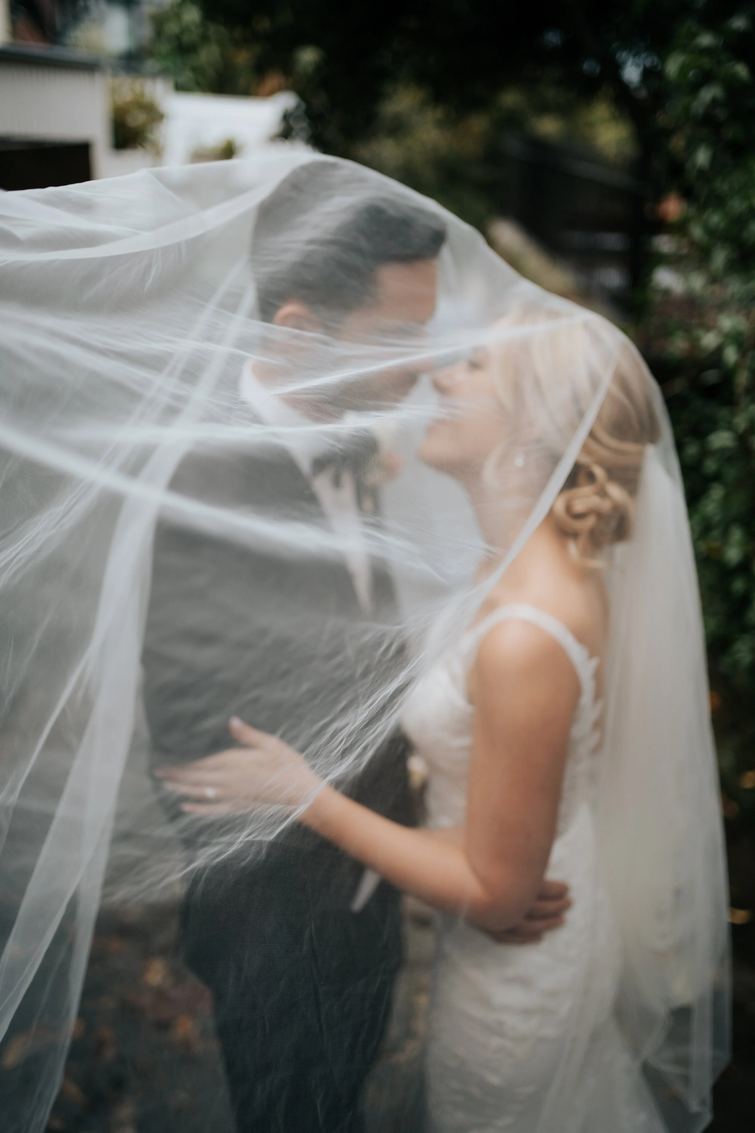 A bride and groom are close together under a sheer veil, sharing a kiss outdoors.
