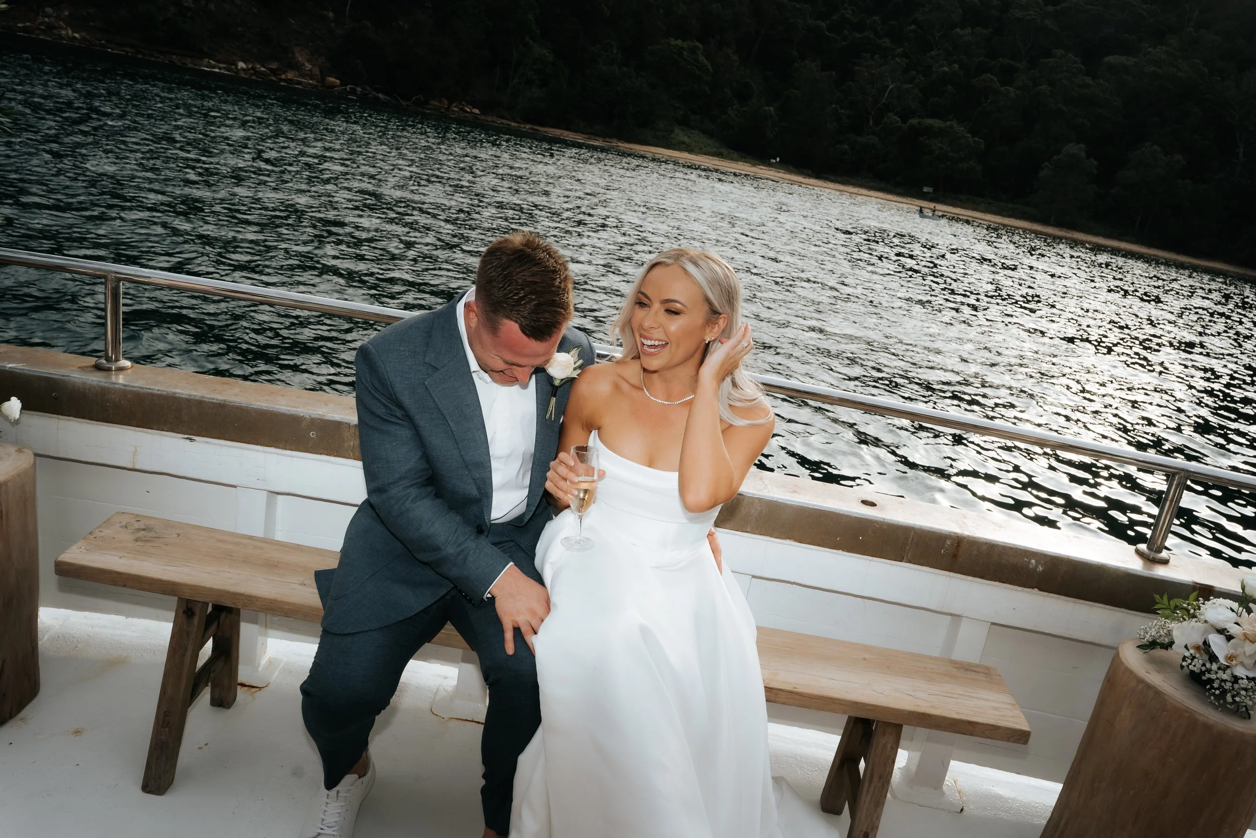 A newlywed couple on a boat, laughing and enjoying champagne, with water and trees in the background.