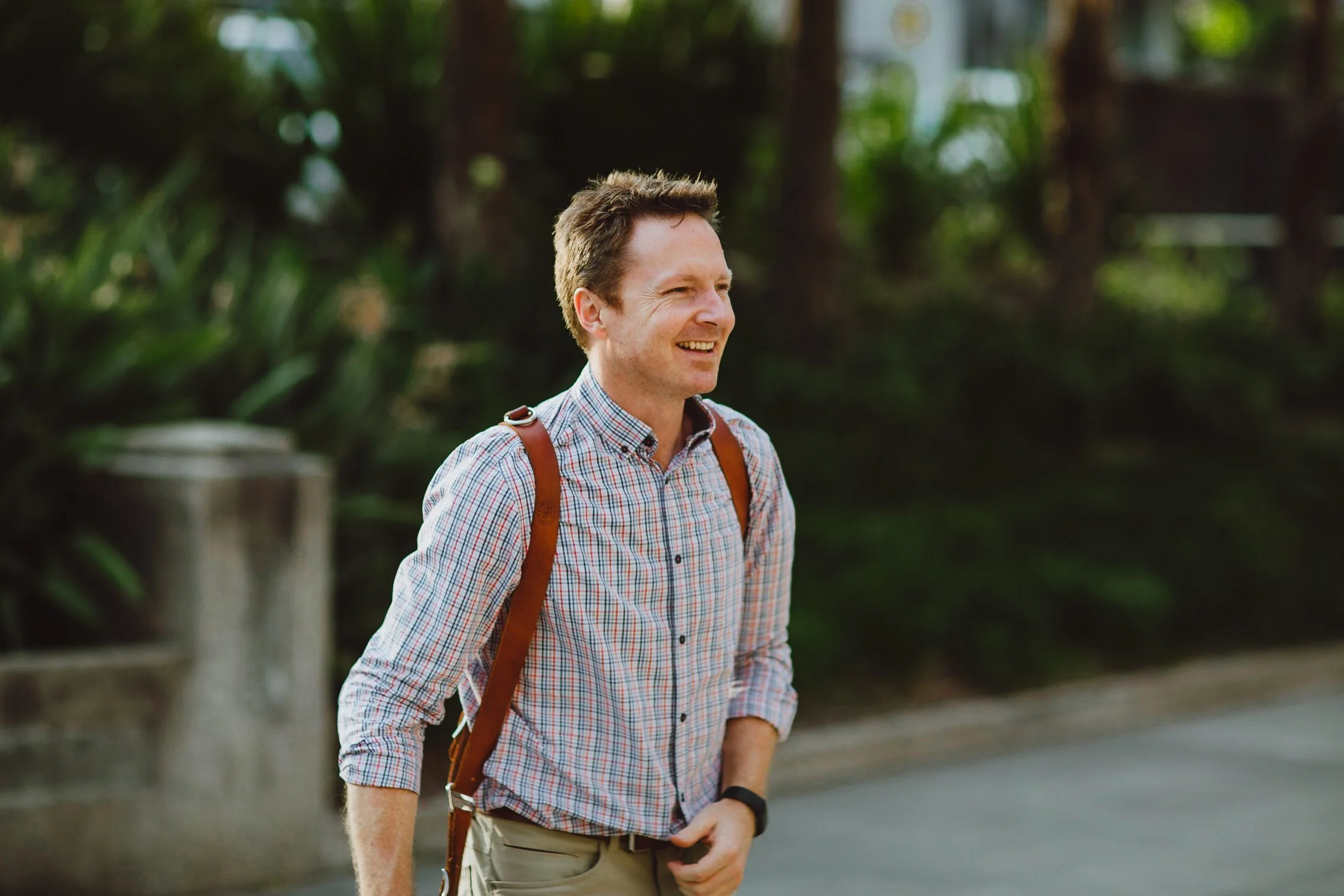 A smiling man walking outdoors with a backpack on his shoulders, wearing a checkered shirt.