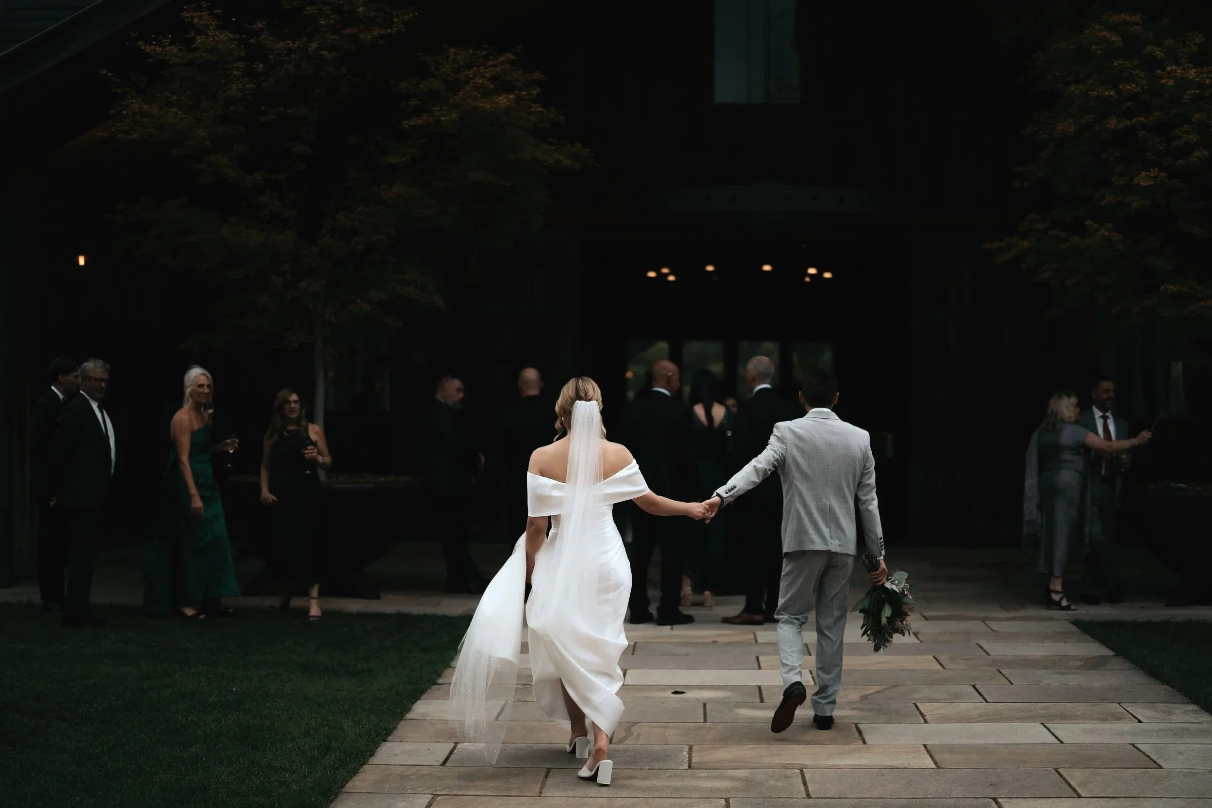A bride and groom holding hands walking away from guests at a wedding ceremony outside in the evening.