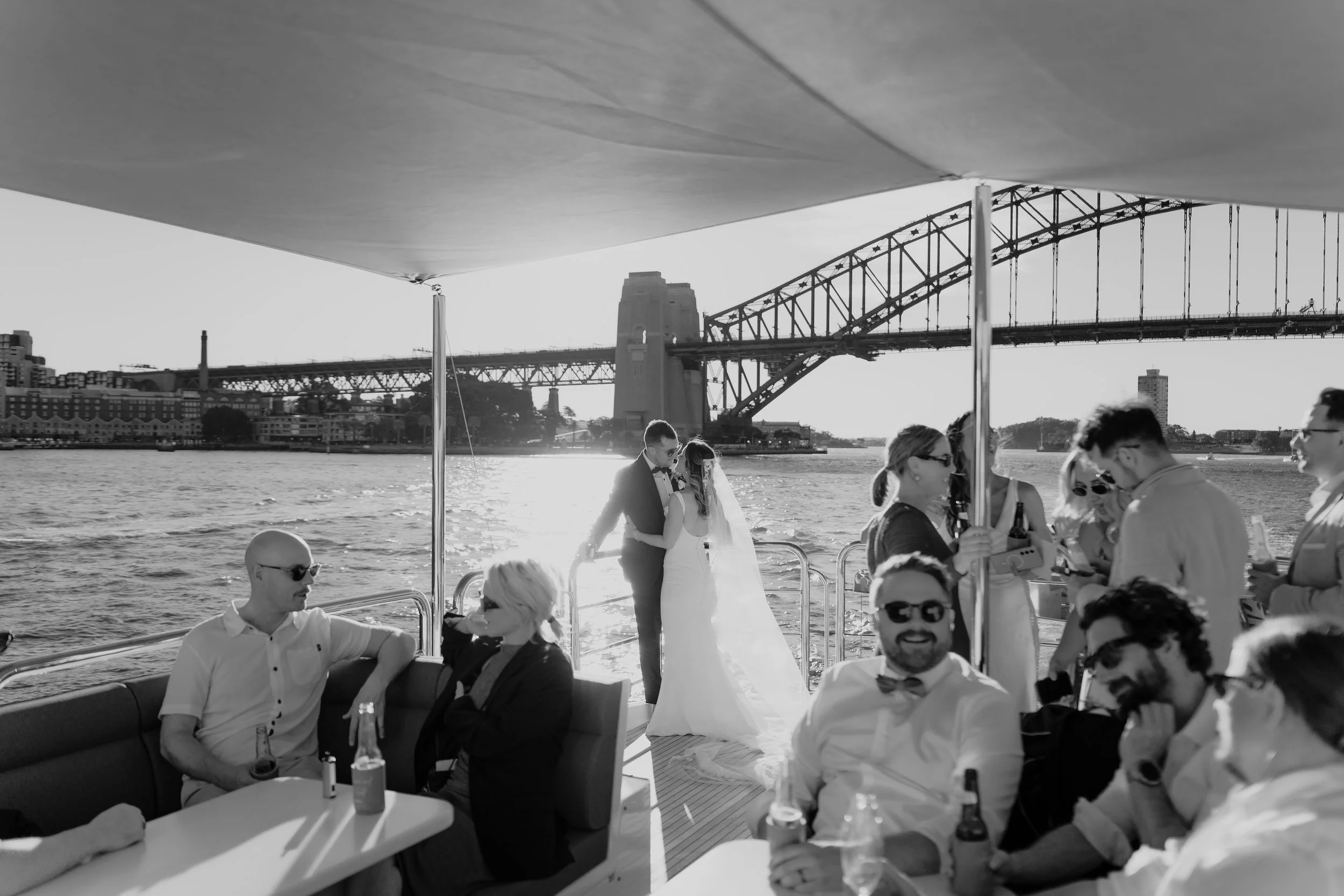 A bride and groom kissing on the deck of a boat with Sydney Harbour Bridge in the background, surrounded by wedding guests during sunset.