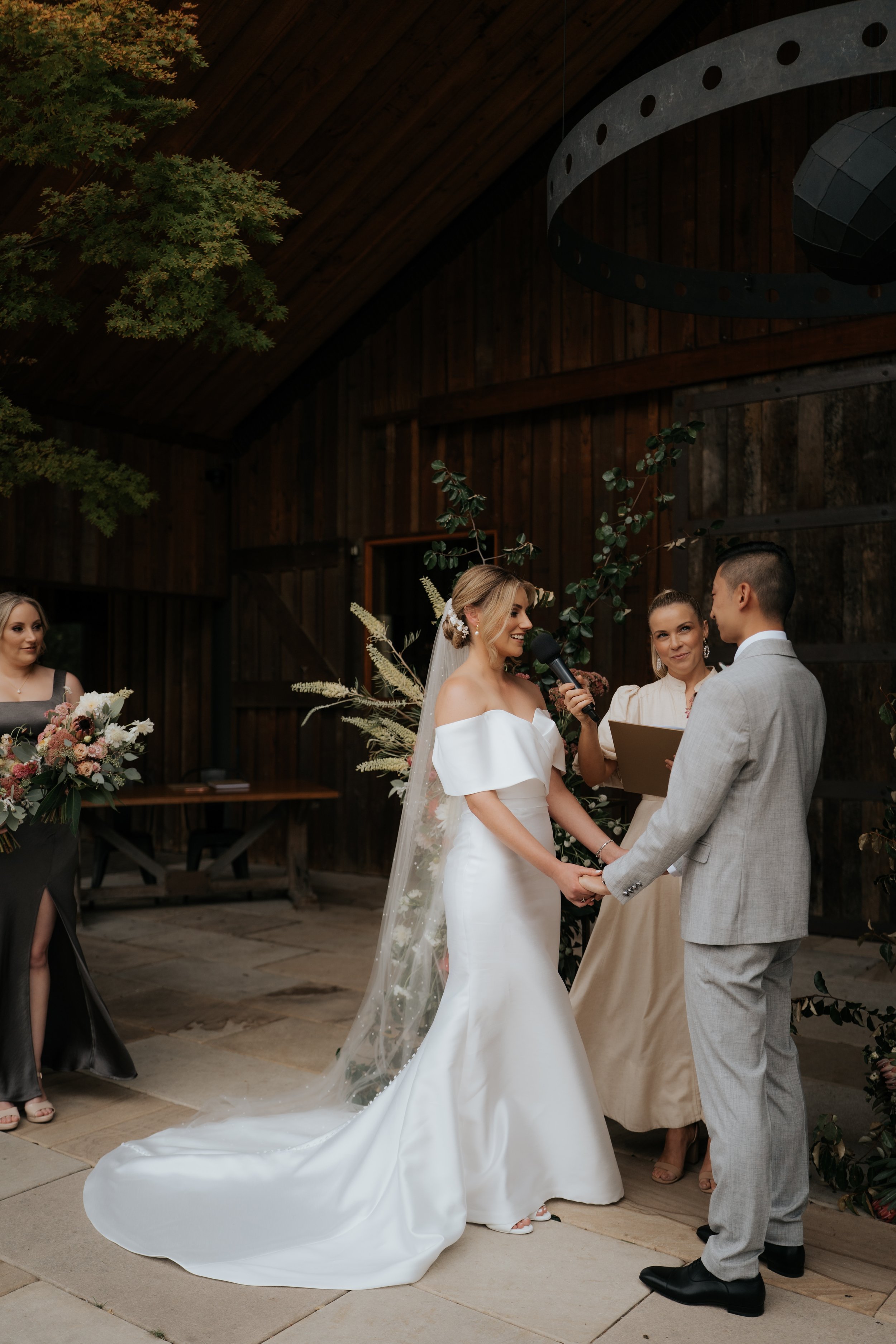 A bride and groom holding hands during their wedding ceremony inside a rustic wooden venue, with a female officiant and a bridesmaid holding a bouquet nearby.