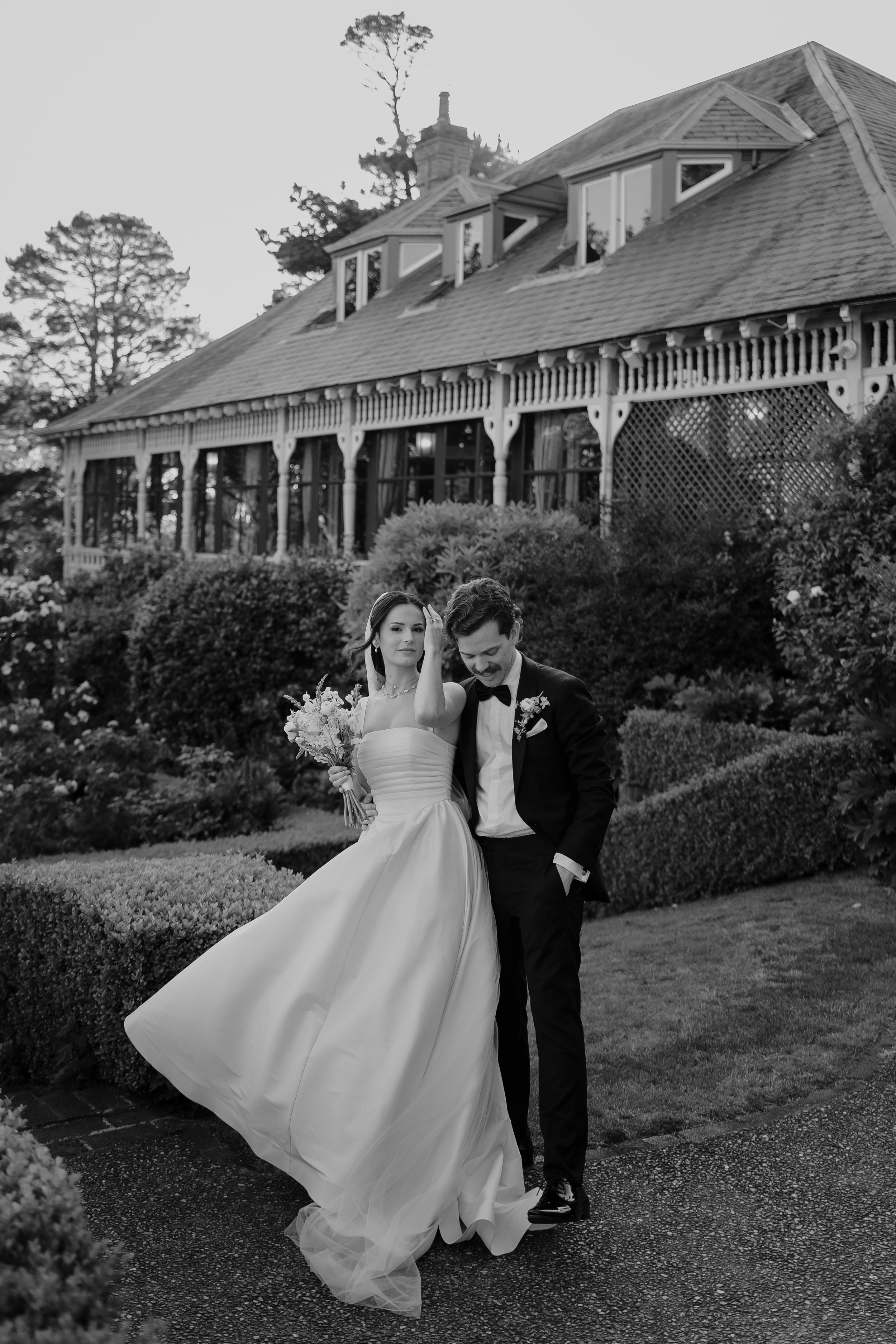 Black and white photo of a bride and groom walking on a garden path, with the bride holding a bouquet and wearing a strapless wedding dress, and the groom in a tuxedo with a bow tie, in front of a large house with a porch and surrounding lush gardens.