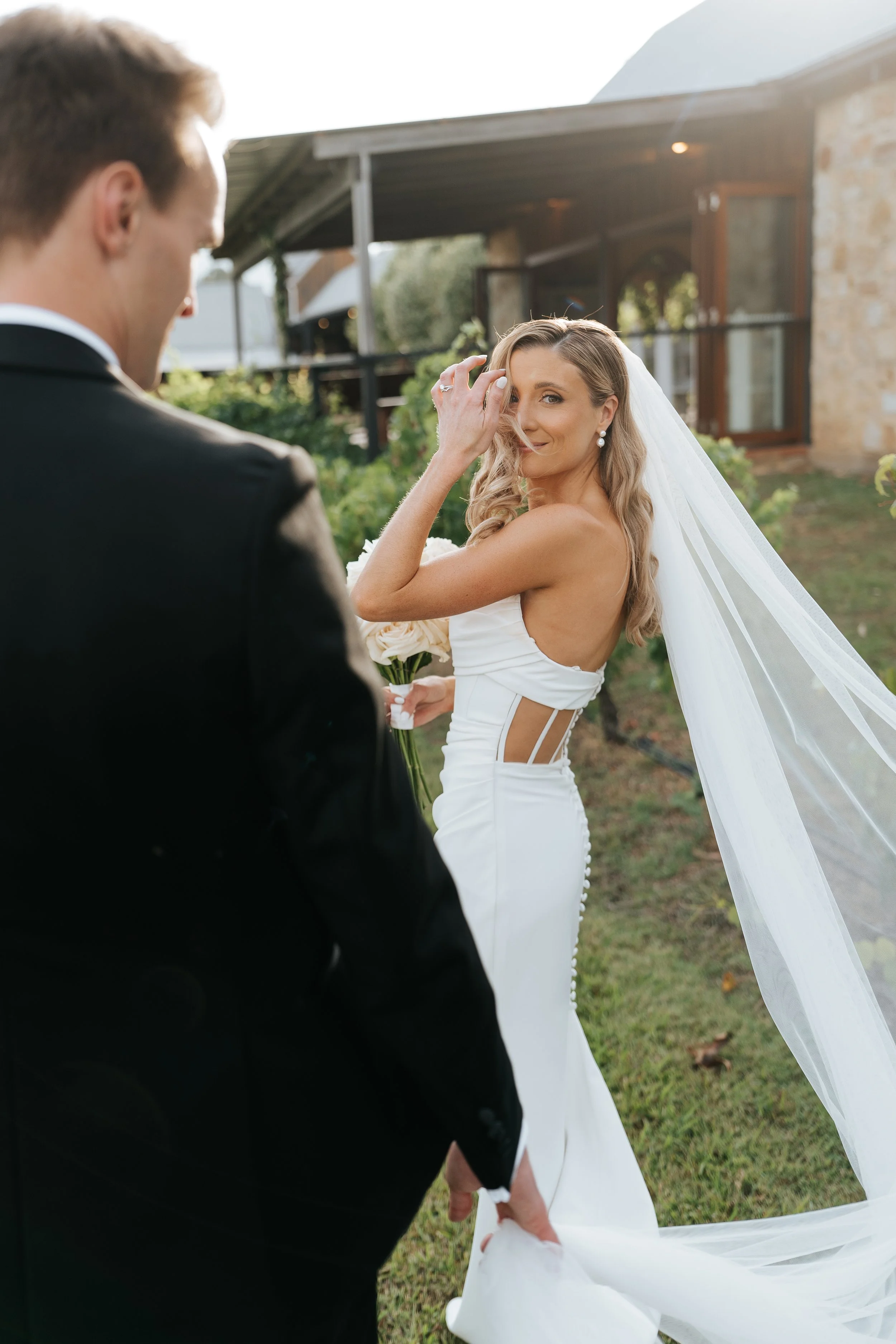 A bride in a strapless white wedding gown with a long veil, holding a bouquet of flowers, stands outdoors near a rustic building. She is smiling and touching her hair, while a groom in a black tuxedo is partially visible in the foreground.