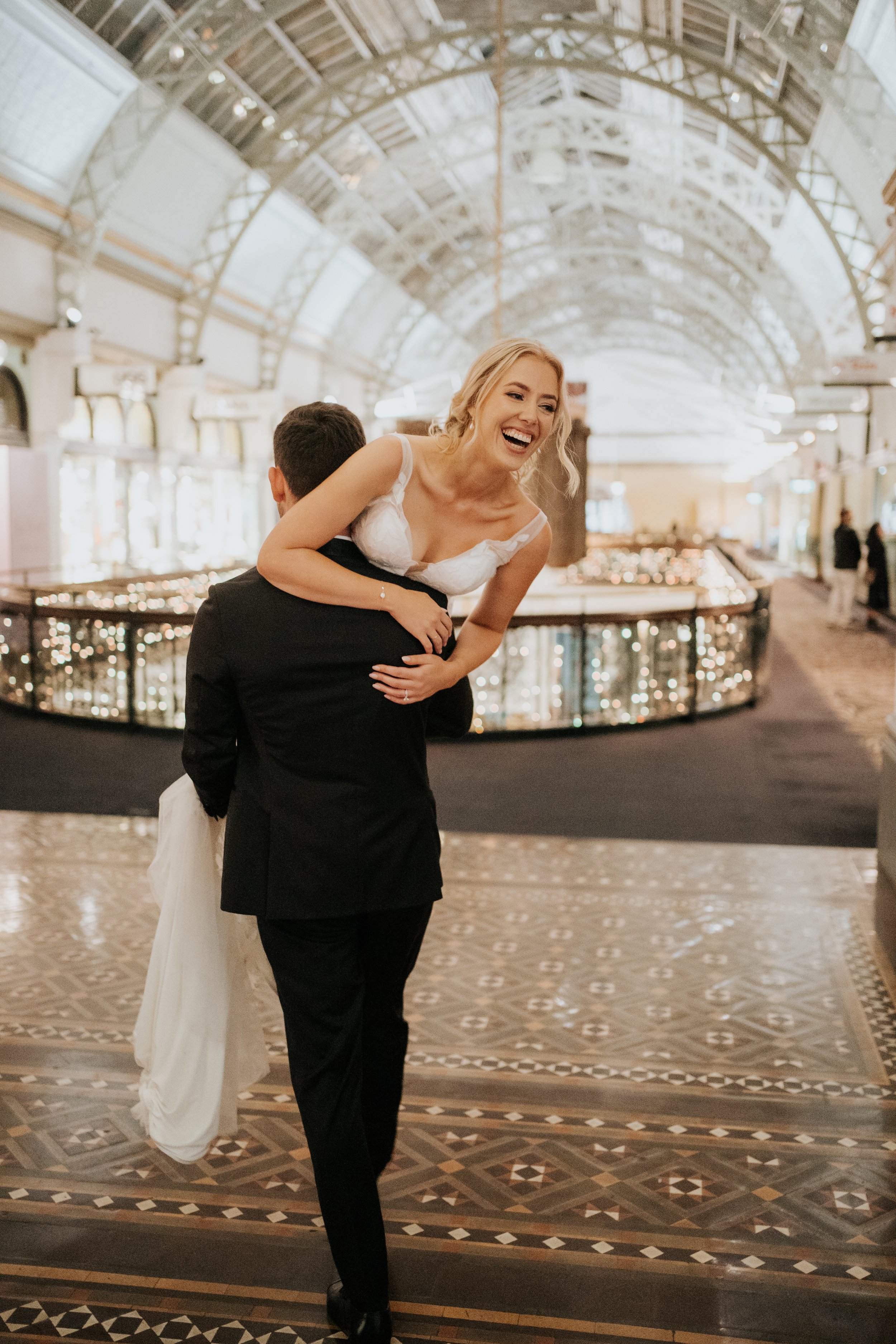 A bridal couple celebrating, with the groom carrying the bride inside an elegant, glass-domed building.