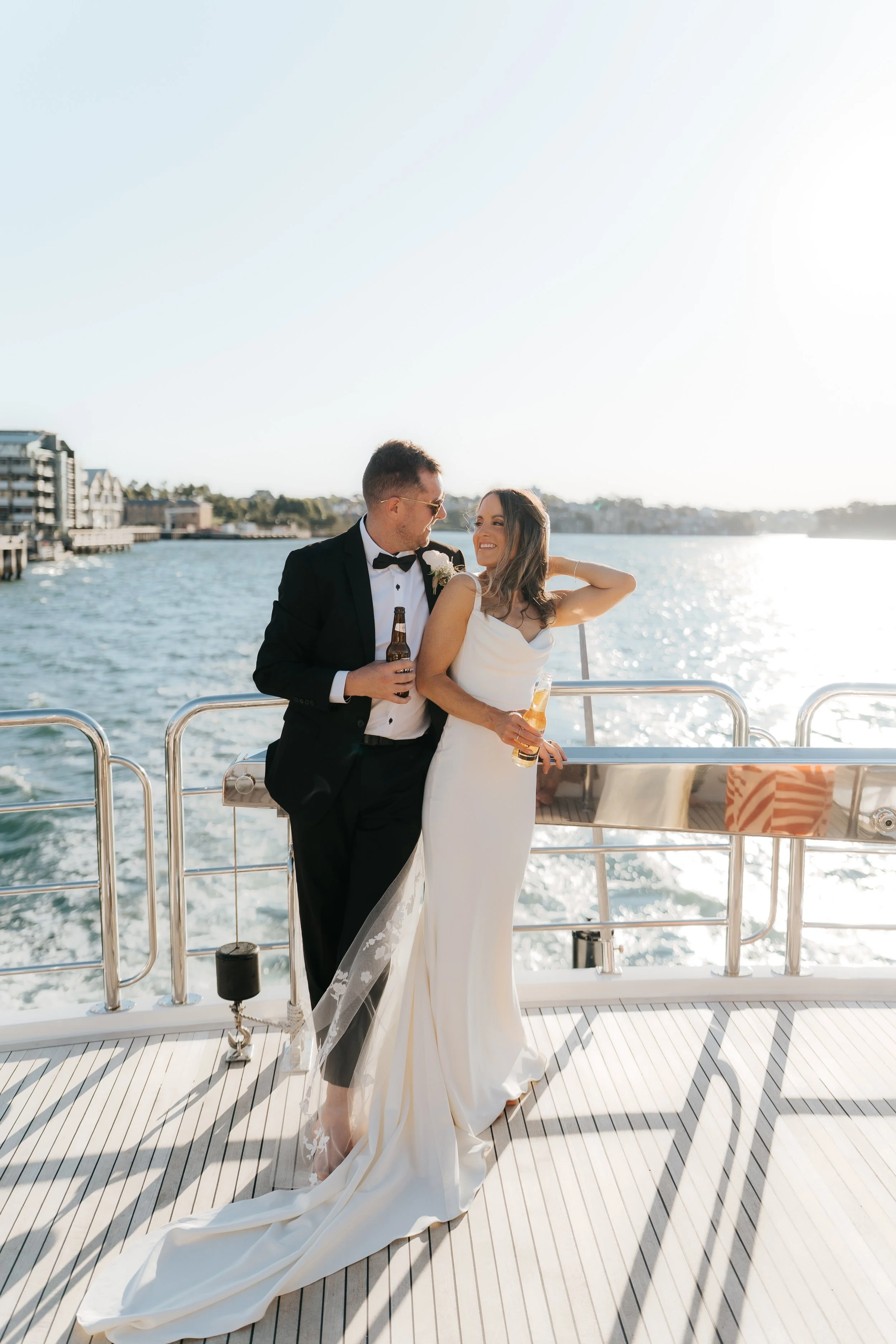 A newlywed couple in formal wedding attire enjoying drinks on a boat with water and buildings in the background, during sunset.