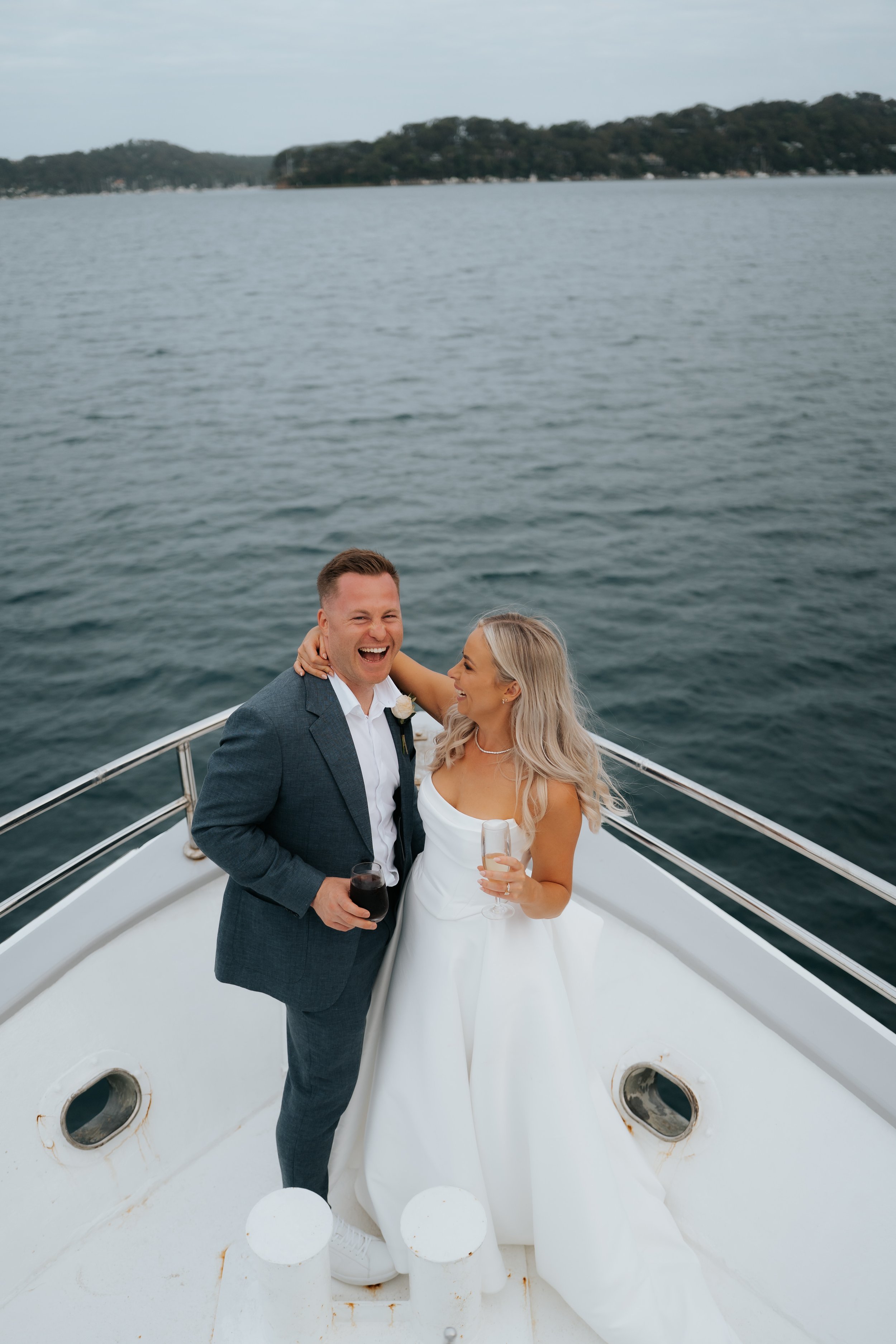 A newlywed couple celebrating on a yacht, with the man in a gray suit and the woman in a white wedding dress, holding wine glasses, smiling and embracing.