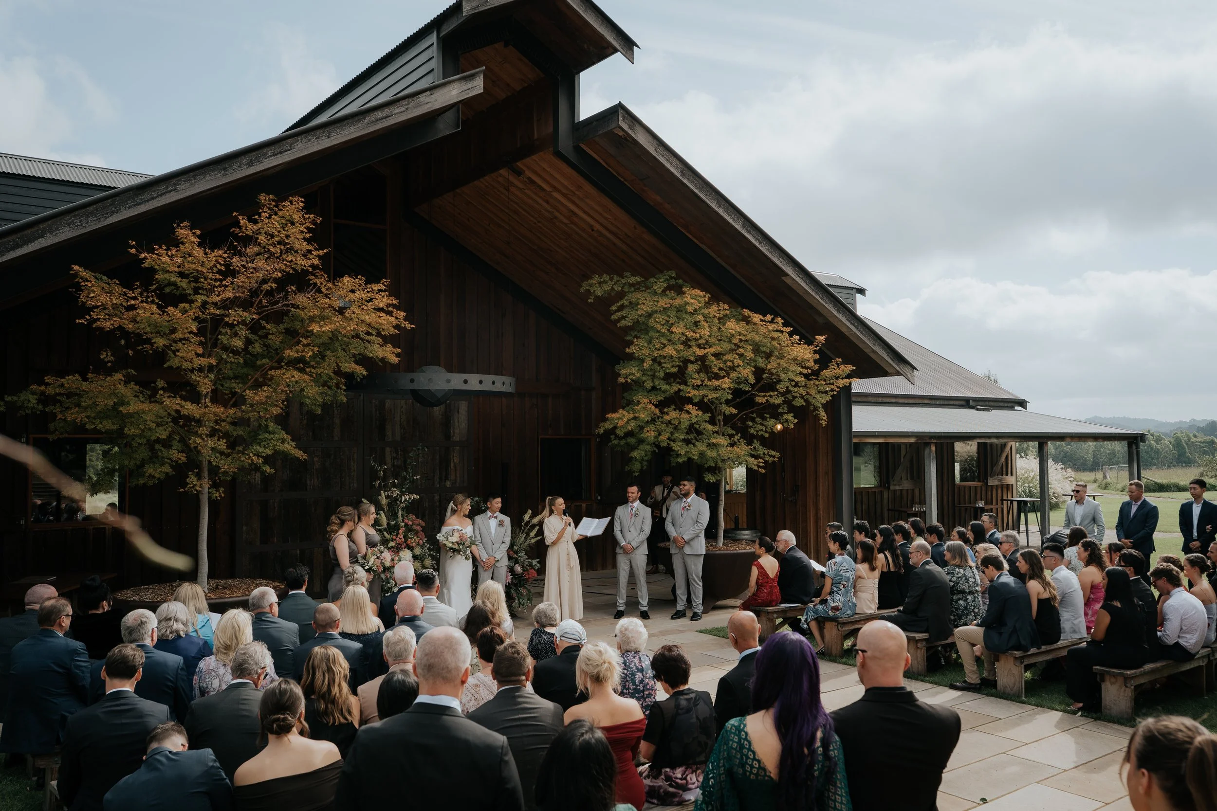 Wedding ceremony taking place outdoors at a rustic barn venue with guests seated on benches and a couple at the altar.