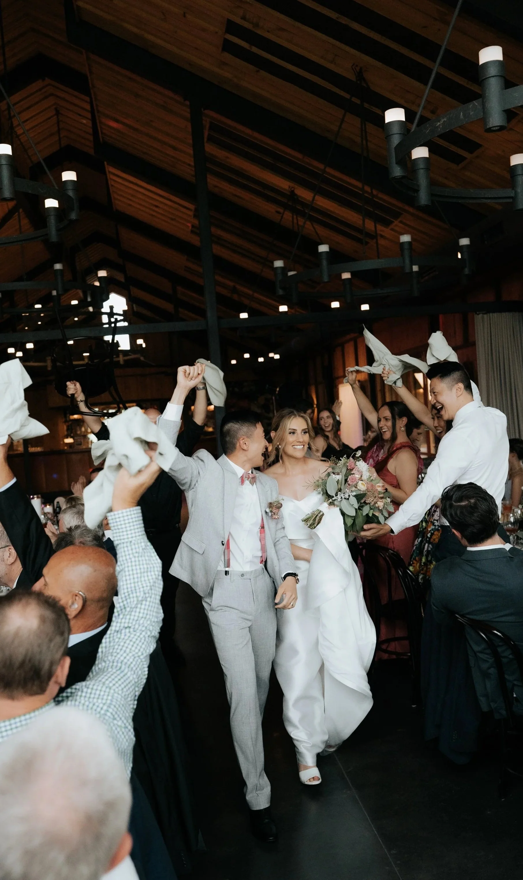 A bride and groom celebrating at their wedding reception, with guests cheering and waving napkins, in a warmly lit wood-paneled venue.
