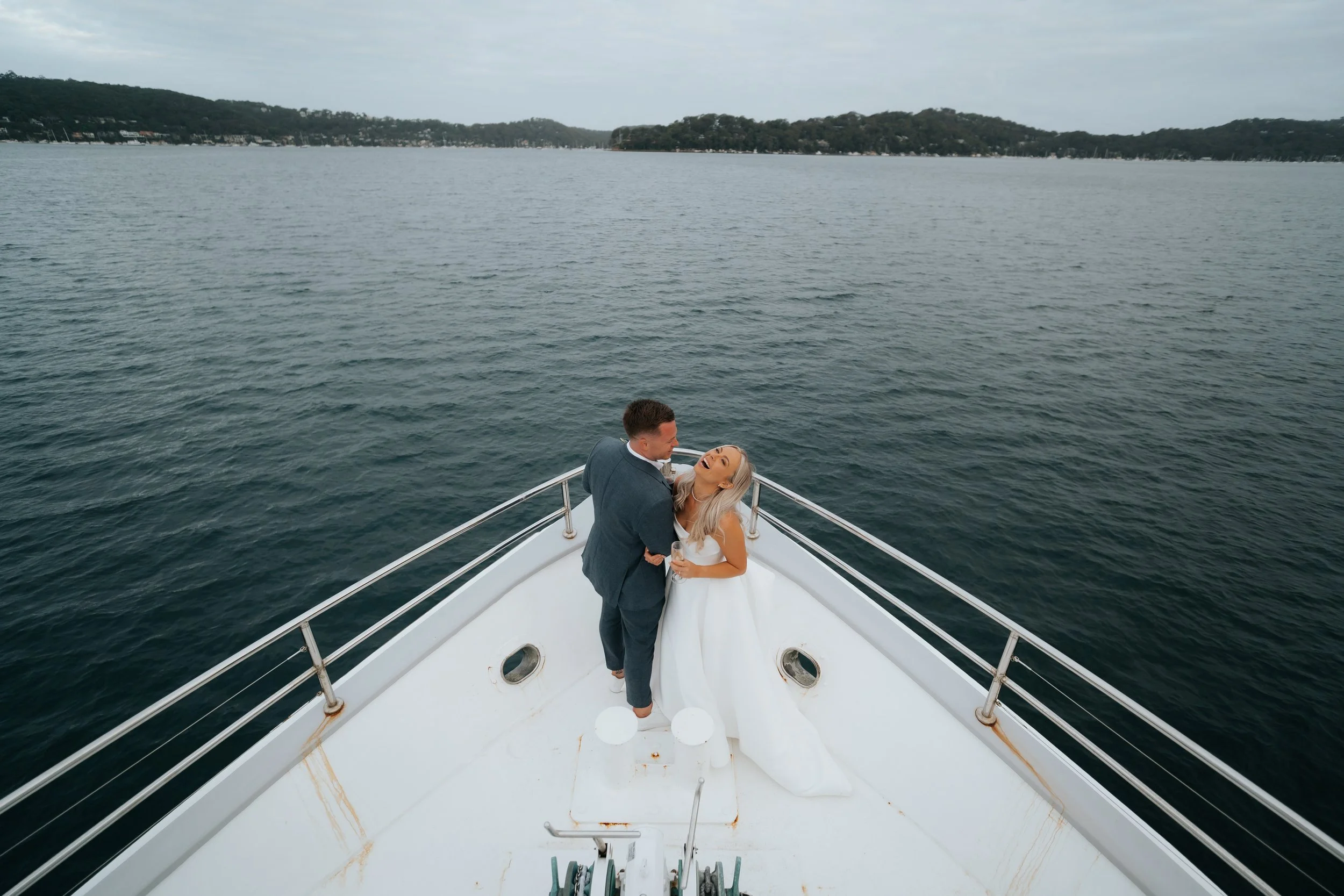 A bride and groom dance and smile on the deck of a boat on a calm body of water with hills and trees in the background.