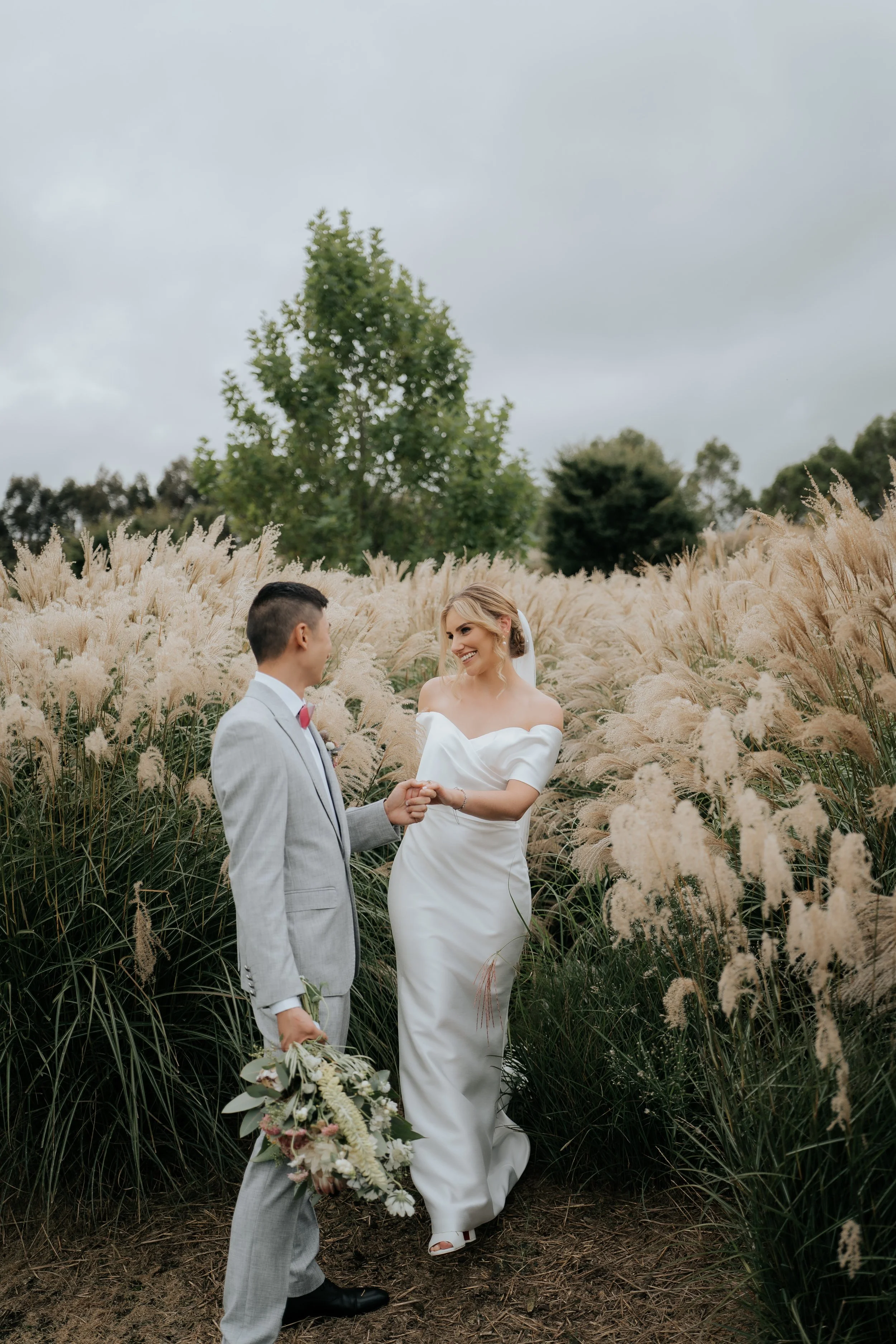 A bride and groom standing in a field of tall, feathery grasses, holding hands and smiling at each other during their wedding photoshoot.