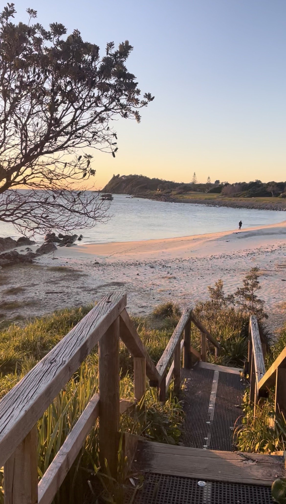 A set of wooden stairs leading down to a beach with white sand, trees hstailing, and a person walking along the shoreline during sunset.