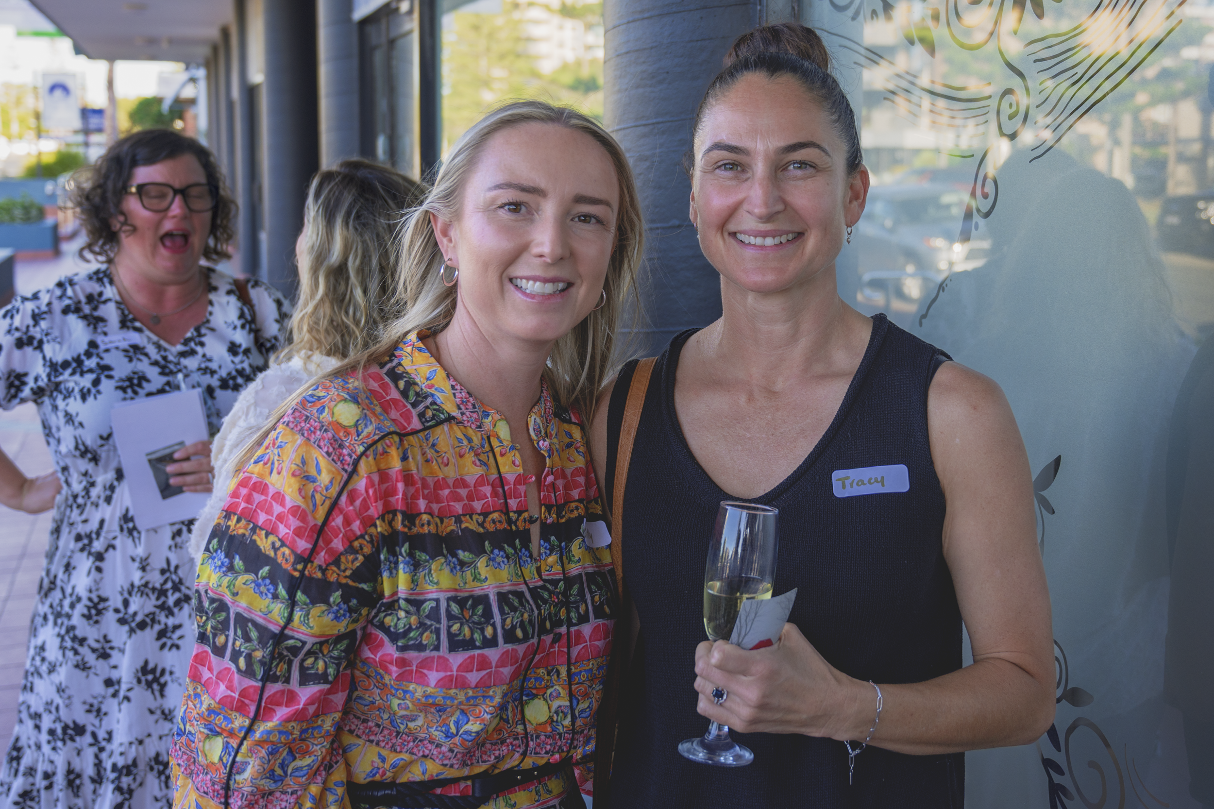 Two women smiling and posing for a photo at an outdoor event, one holding a glass of champagne, with people in the background