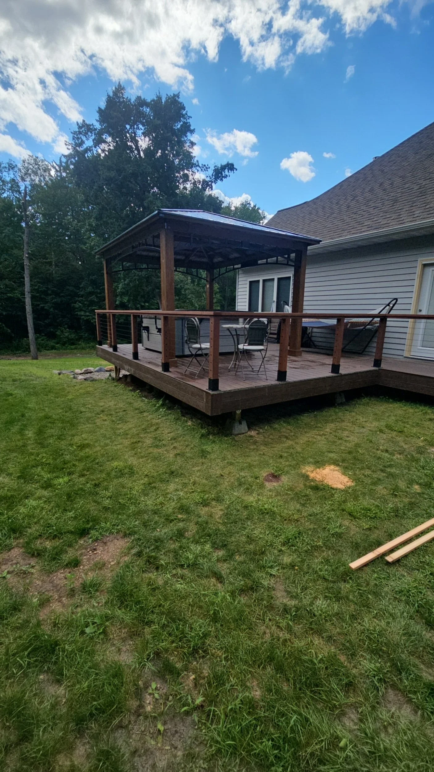 A backyard wooden deck attached to a house, featuring a covered gazebo, patio furniture, and a cooler, surrounded by green grass and trees.