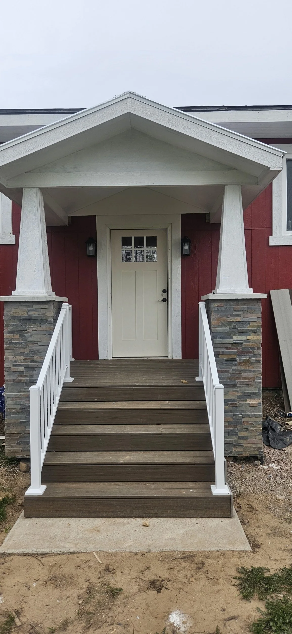Front porch of a house with wooden stairs, white railing, stone pillars, and a beige door under a white gabled roof.