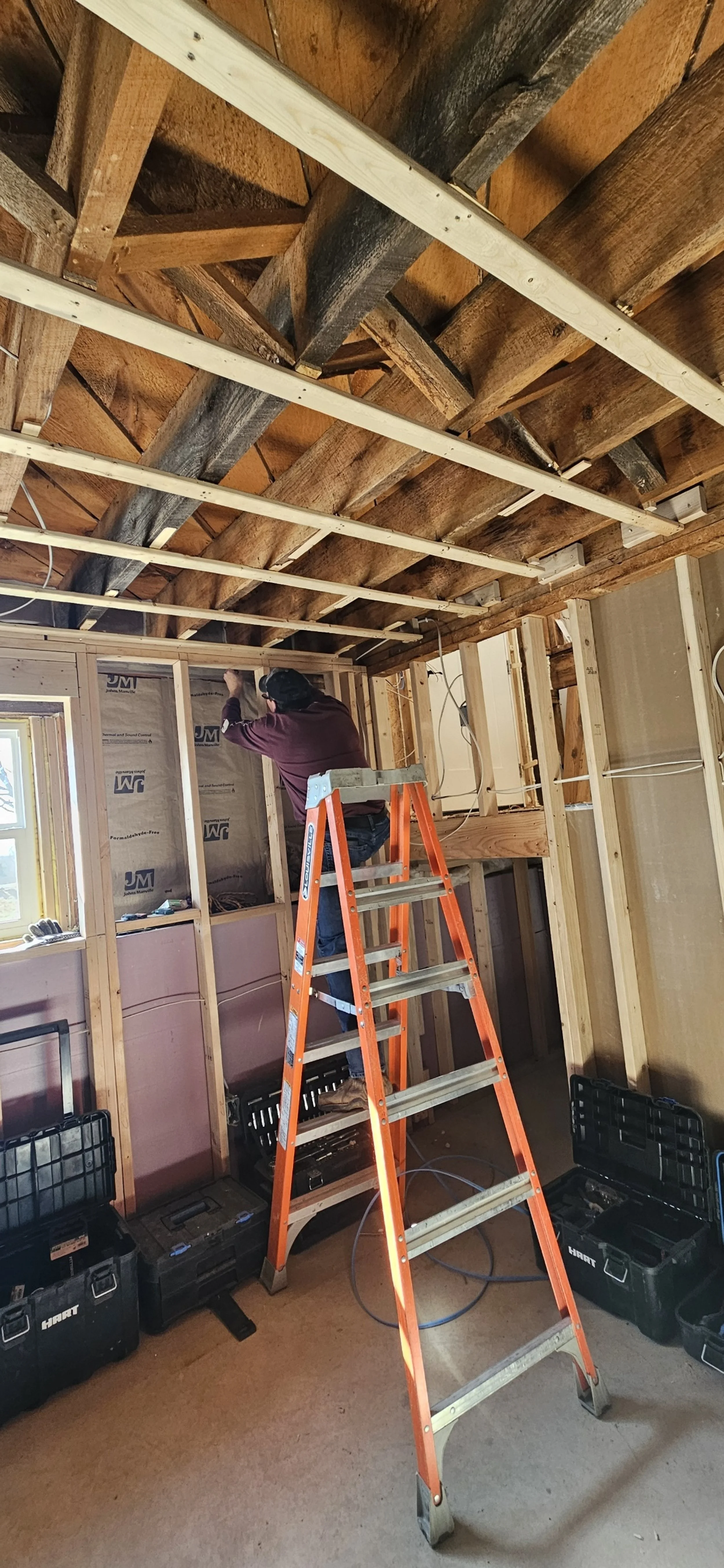 A person working on electrical wiring inside a partially constructed room with exposed studs and ceiling beams; the person is on an orange ladder near a window with tools and toolboxes on the floor.
