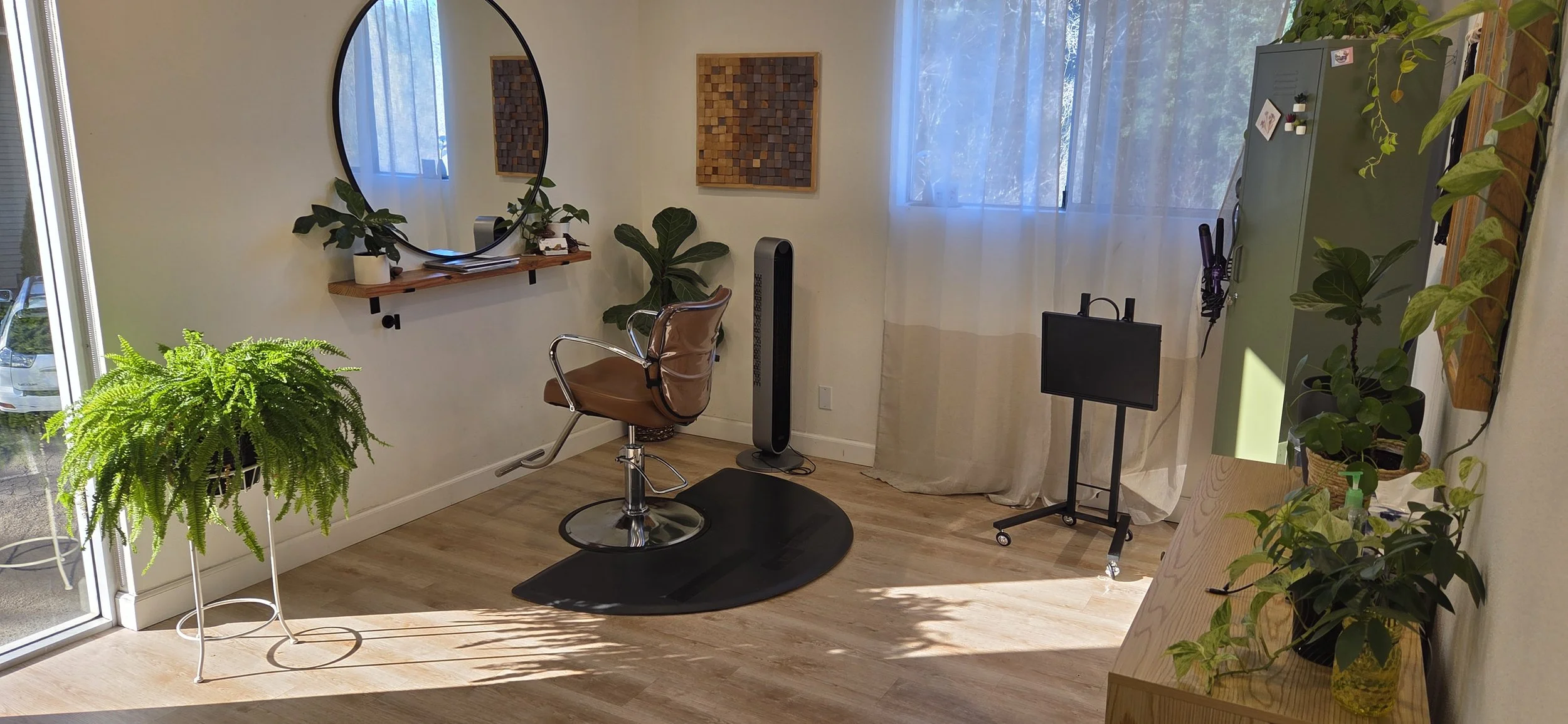 A cozy hair salon corner with a styling chair, a large mirror, and green plants on shelves and stands near a window with sheer curtains, natural sunlight illuminating the space.