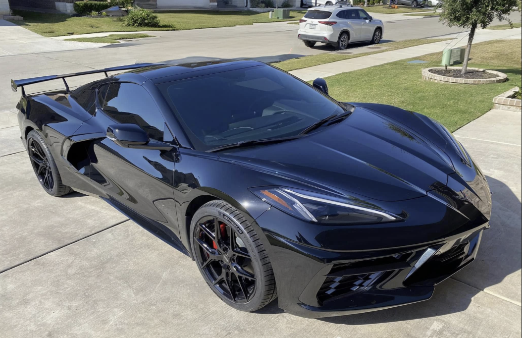 Black sports car parked in driveway on a sunny day with a house, trees, and trash bin in the background.