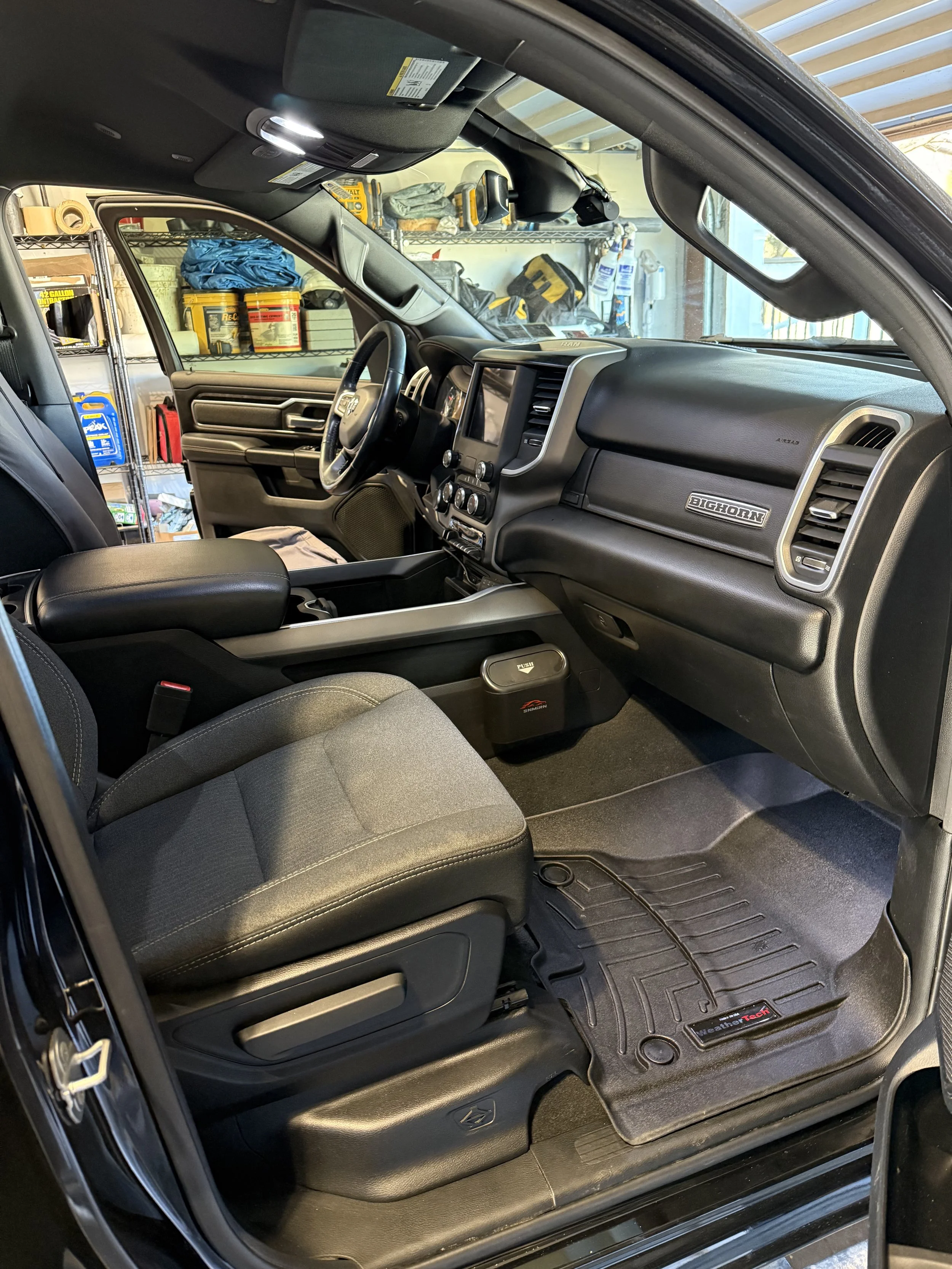 Interior of a pickup truck with a black dashboard, steering wheel, gray fabric seats, and a rubber floor mat, parked in a garage with shelves and tools in the background.