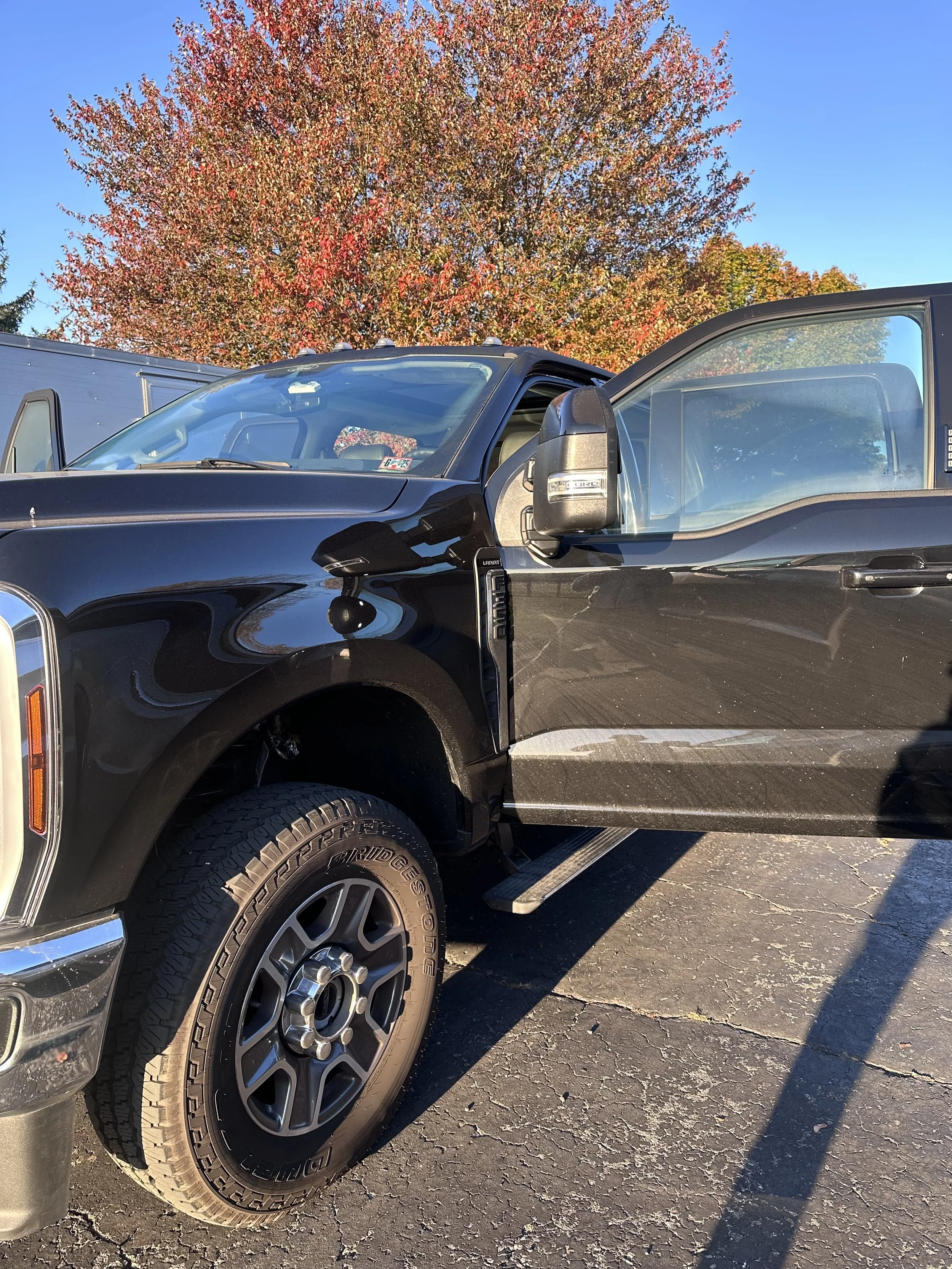 Black pickup truck parked on a paved lot with autumn-colored trees in the background and a clear blue sky.