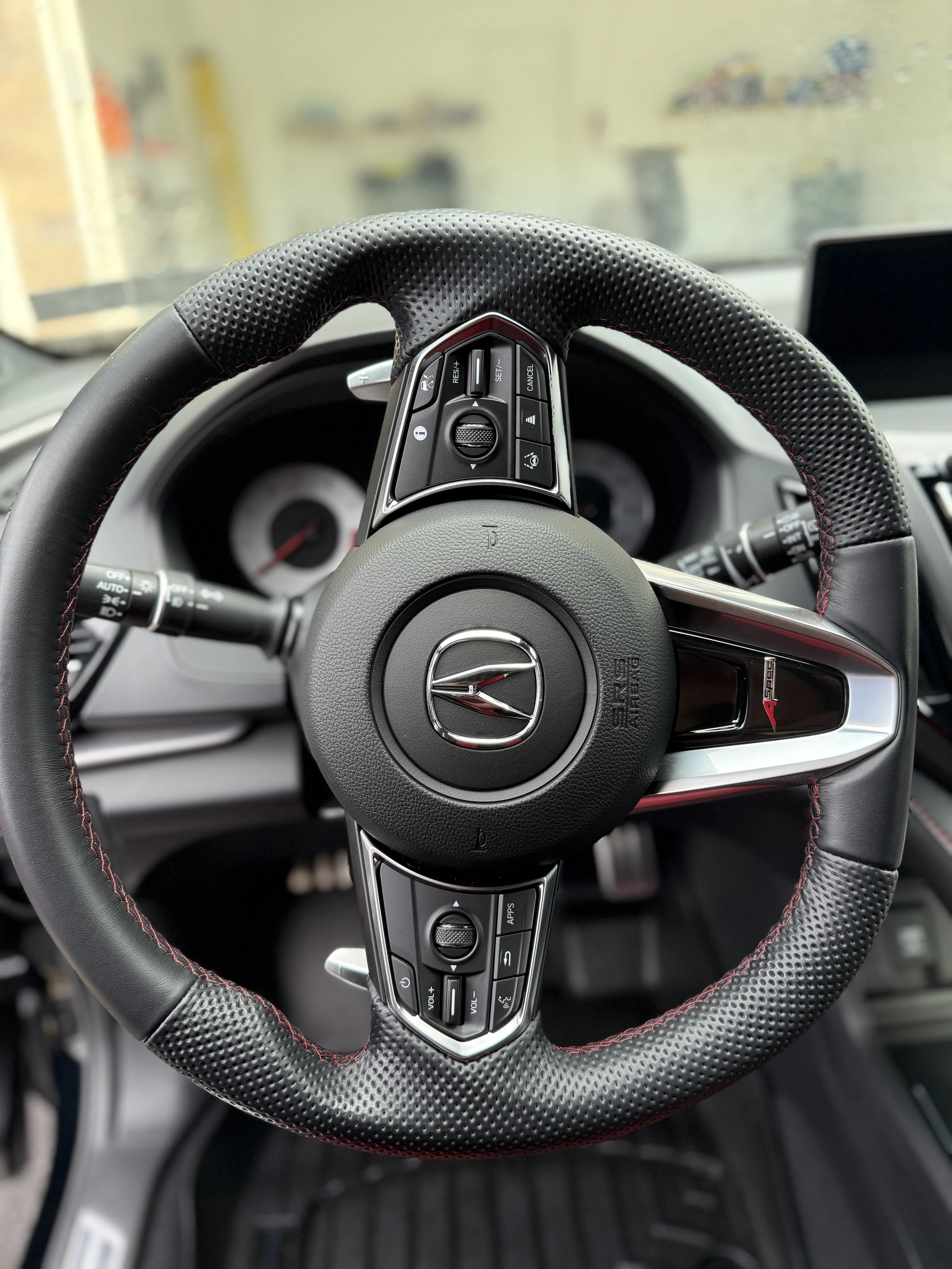 Close-up of a car steering wheel with the Acura logo, featuring mounted control buttons and a perforated leather grip with red stitching.