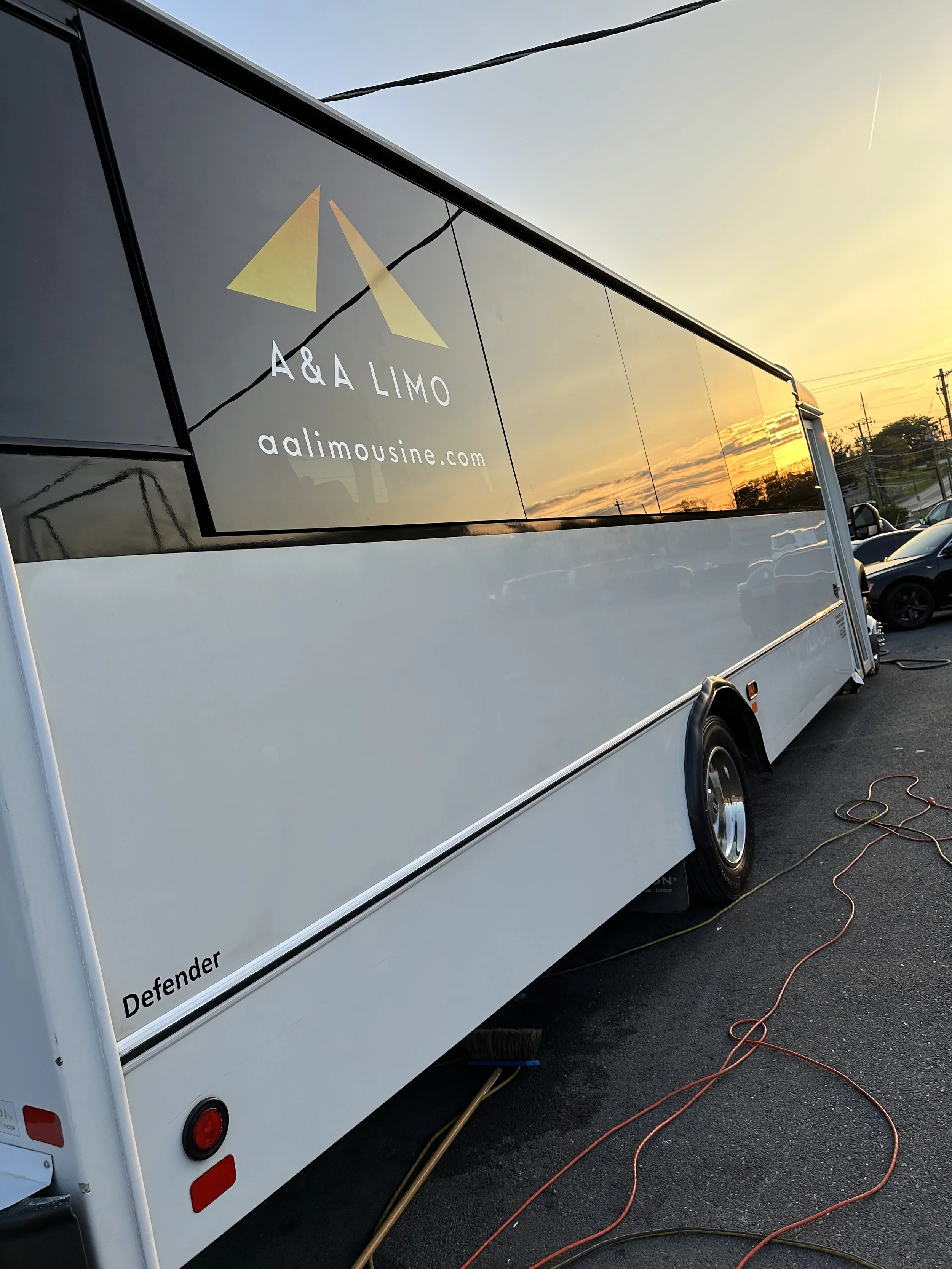 A white catering truck with a black-tinted window reflecting the sunset and power lines. The logo on the window reads 'A & A LIMO' with a website 'aalimousine.com'. The truck is parked on asphalt with extension cords and a broom lying on the ground n
