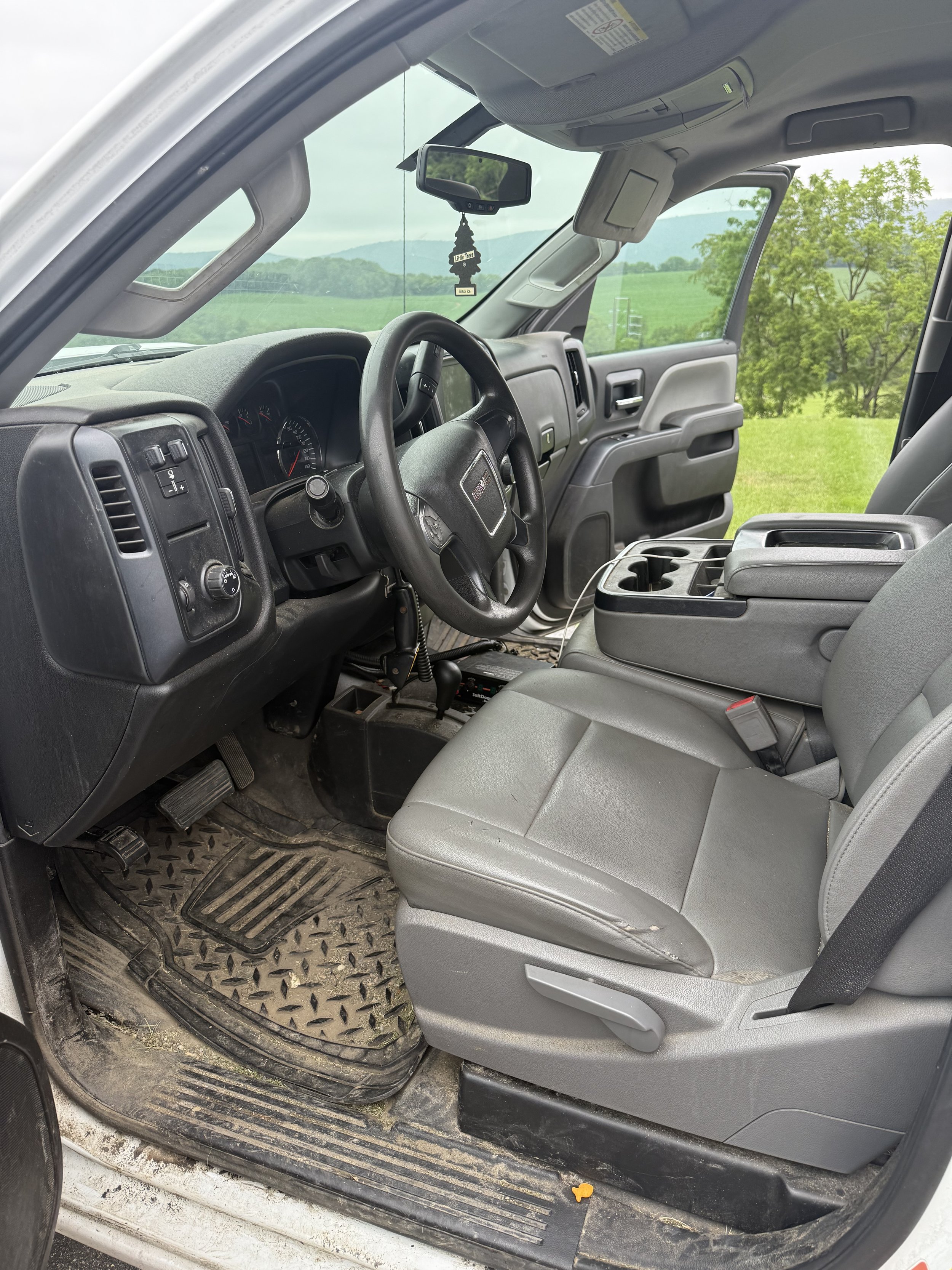 Interior of a gray work van or truck, showing the driver's seat, steering wheel, dashboard, and center console, with a scenic grassy landscape visible through the windows.
