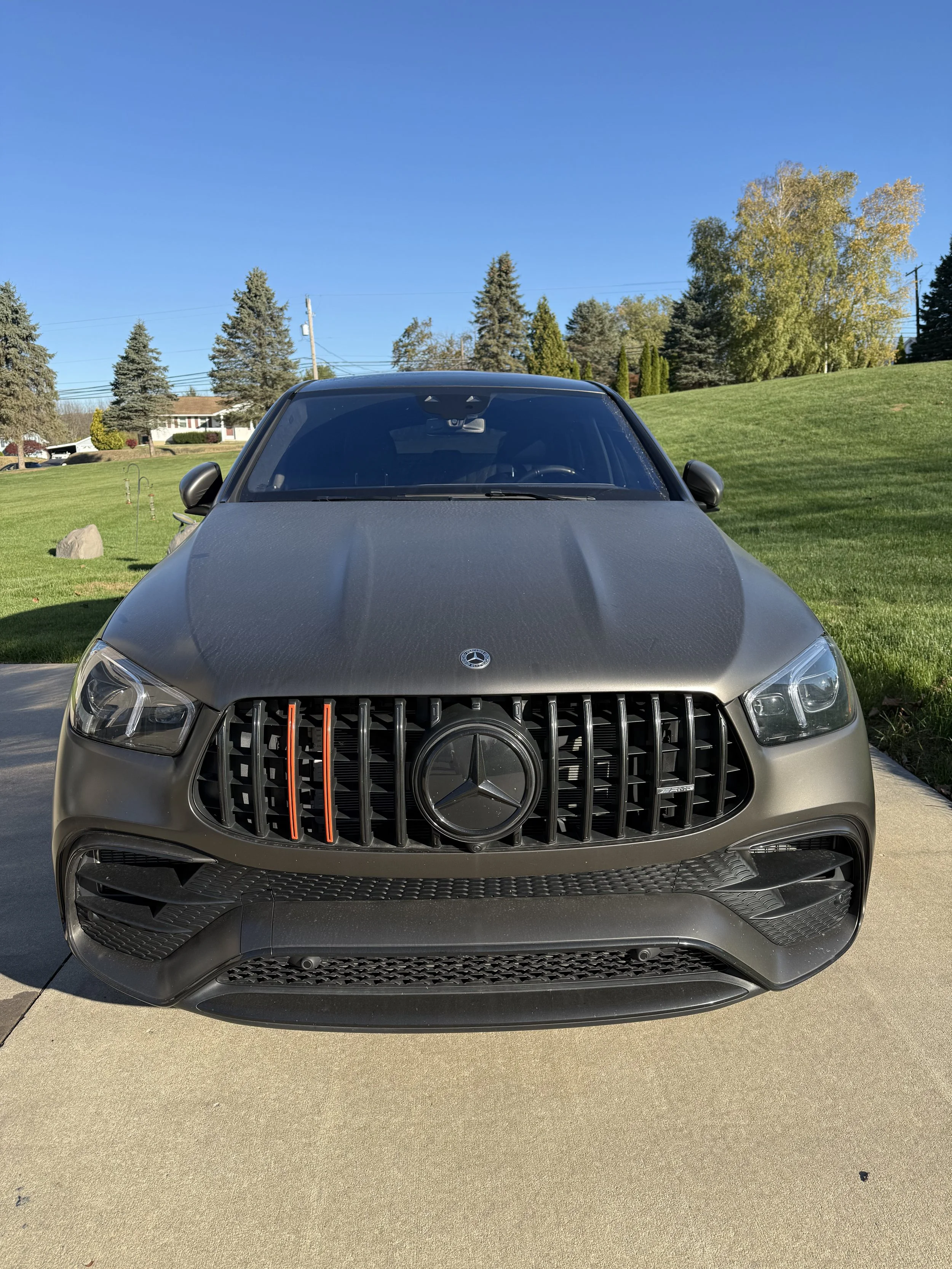 Front view of a black Mercedes-Benz SUV with a matte finish, parked on a driveway in a suburban neighborhood, with grass, trees, and houses in the background under a clear blue sky.