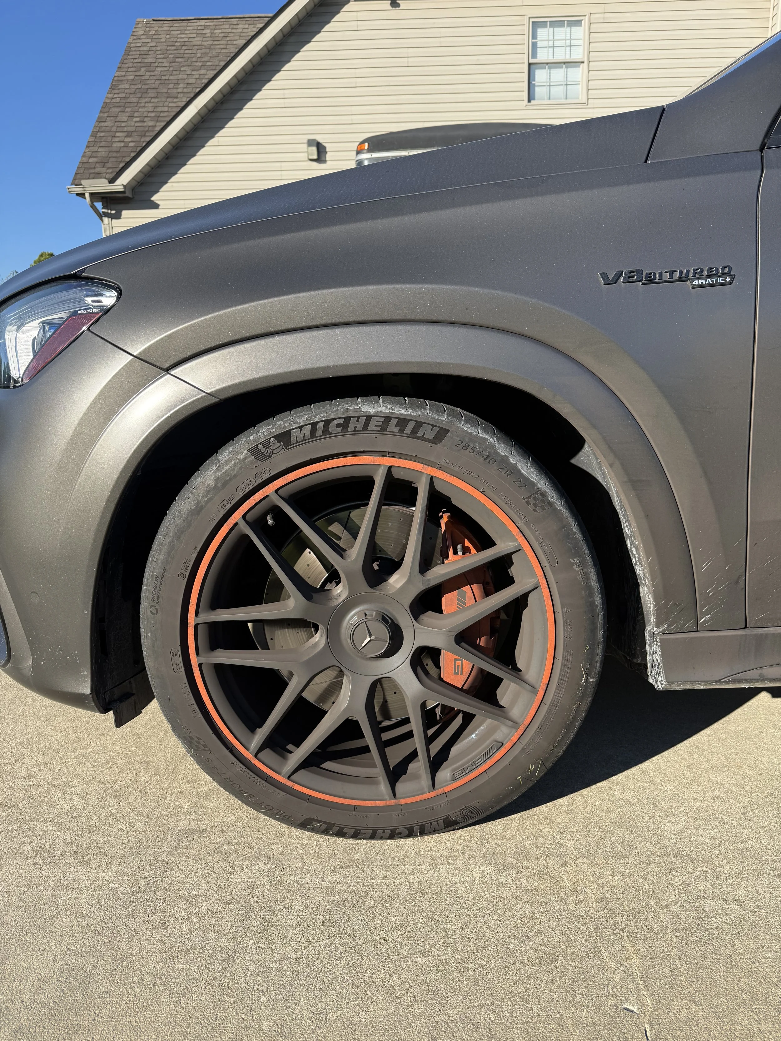 Close-up of a gray Mercedes-Benz car wheel with black rim and orange accents, Michelin tire, and orange brake caliper. Part of the car's front side, including a 'V8 BITURBO' badge, is visible.