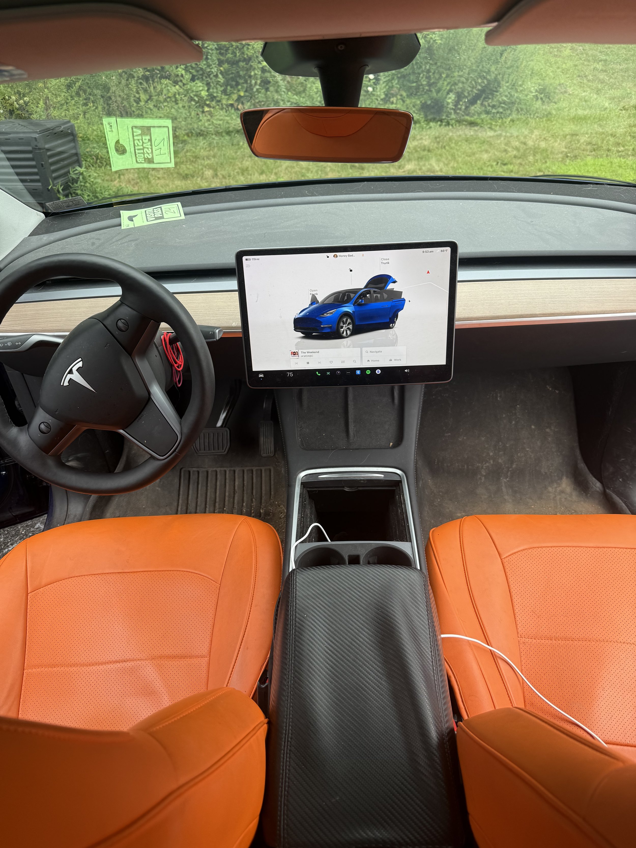 Interior of a Tesla vehicle with orange seats, a steering wheel with a Tesla logo, a large central touchscreen displaying a blue car, and an open outdoor view through the windshield showing greenery.