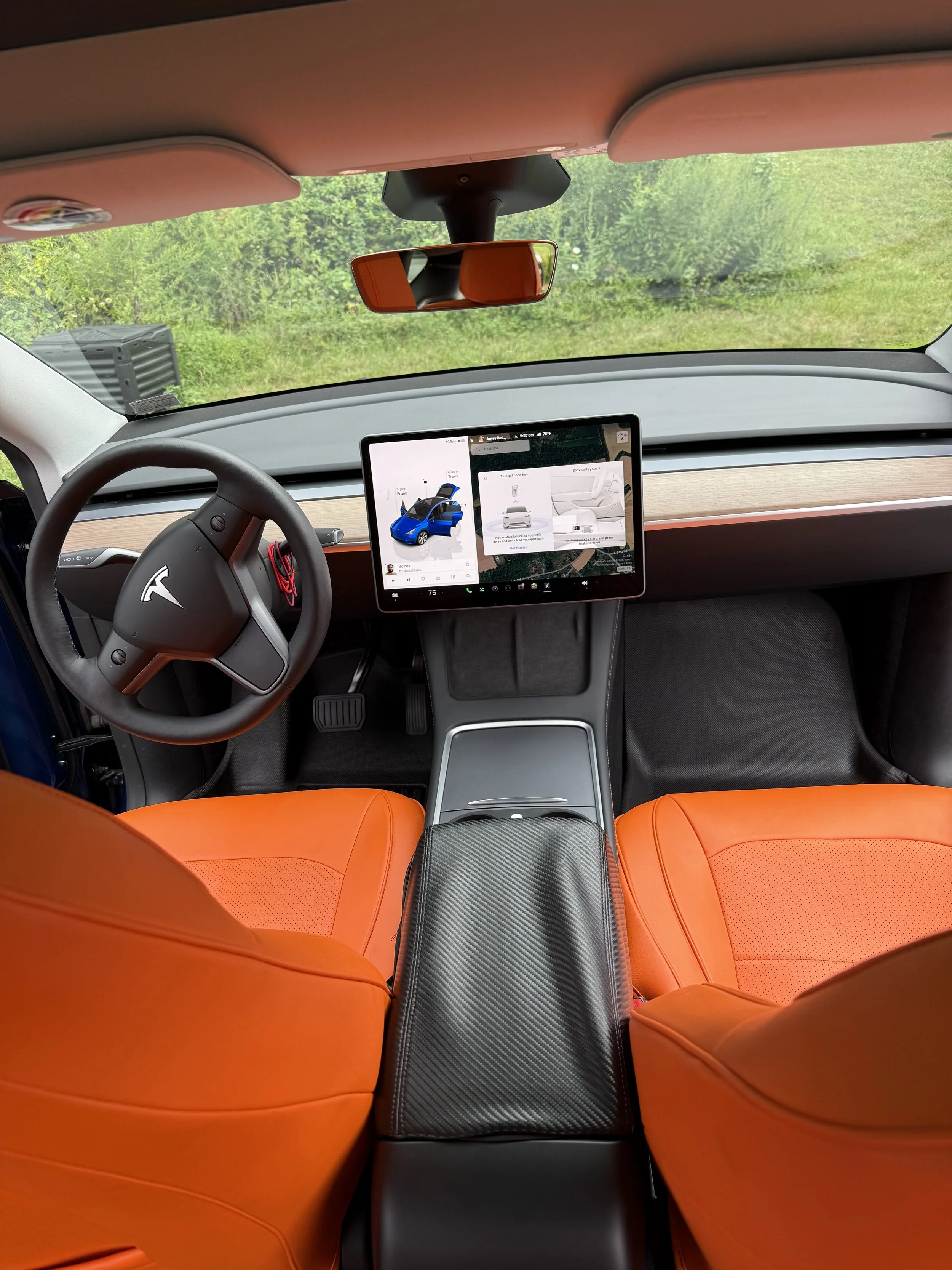 Inside the cockpit of a Tesla vehicle with orange seats, a digital display on the dashboard showing car settings, and a view of green trees through the windshield.