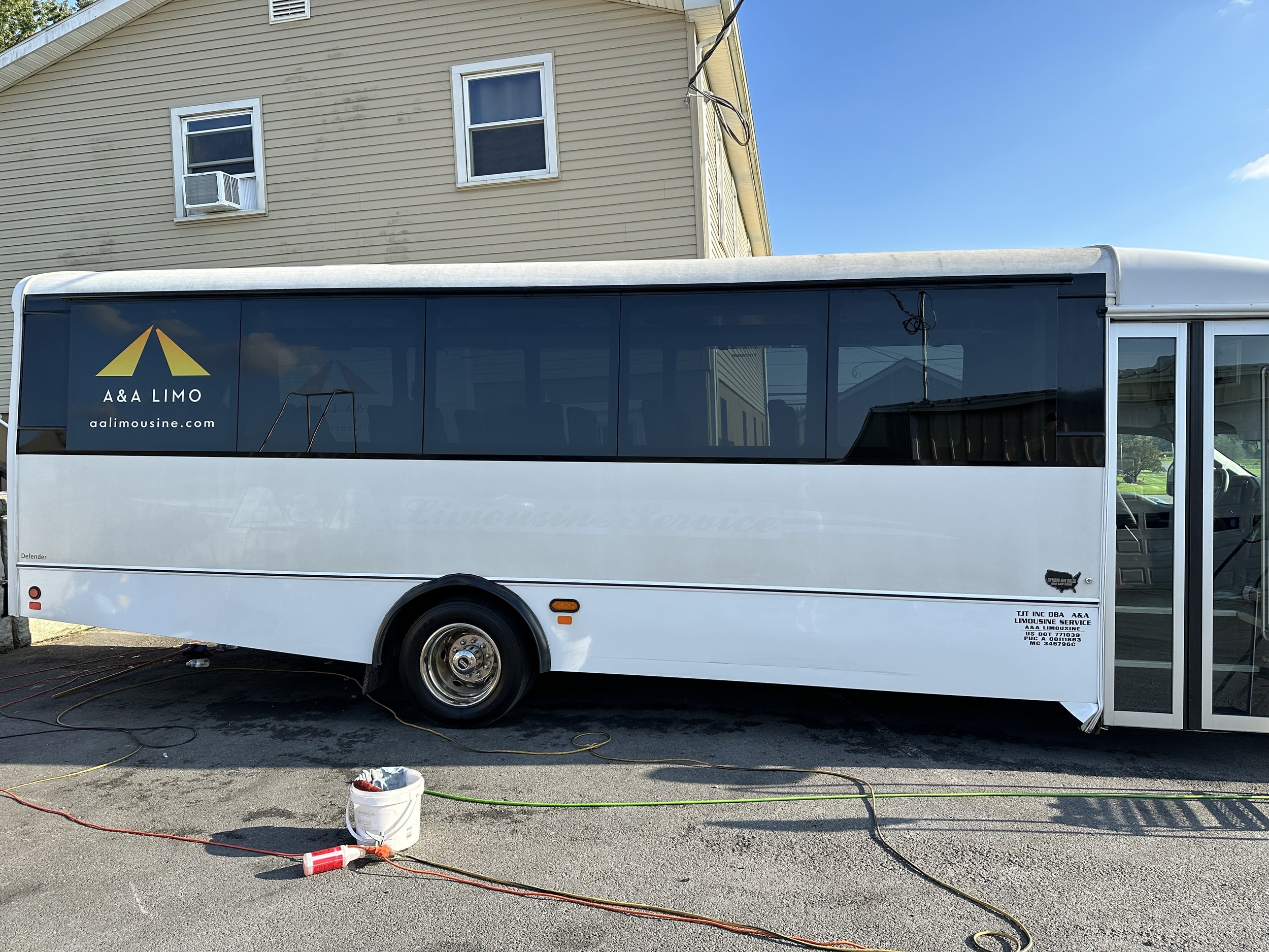 White limousine bus parked outside near a beige house with two windows, one with an air conditioning unit. The bus has the logo 'A&A LIMO' and the website aalimousine.com on the side. There are electrical cords and a white bucket with tools on the gr