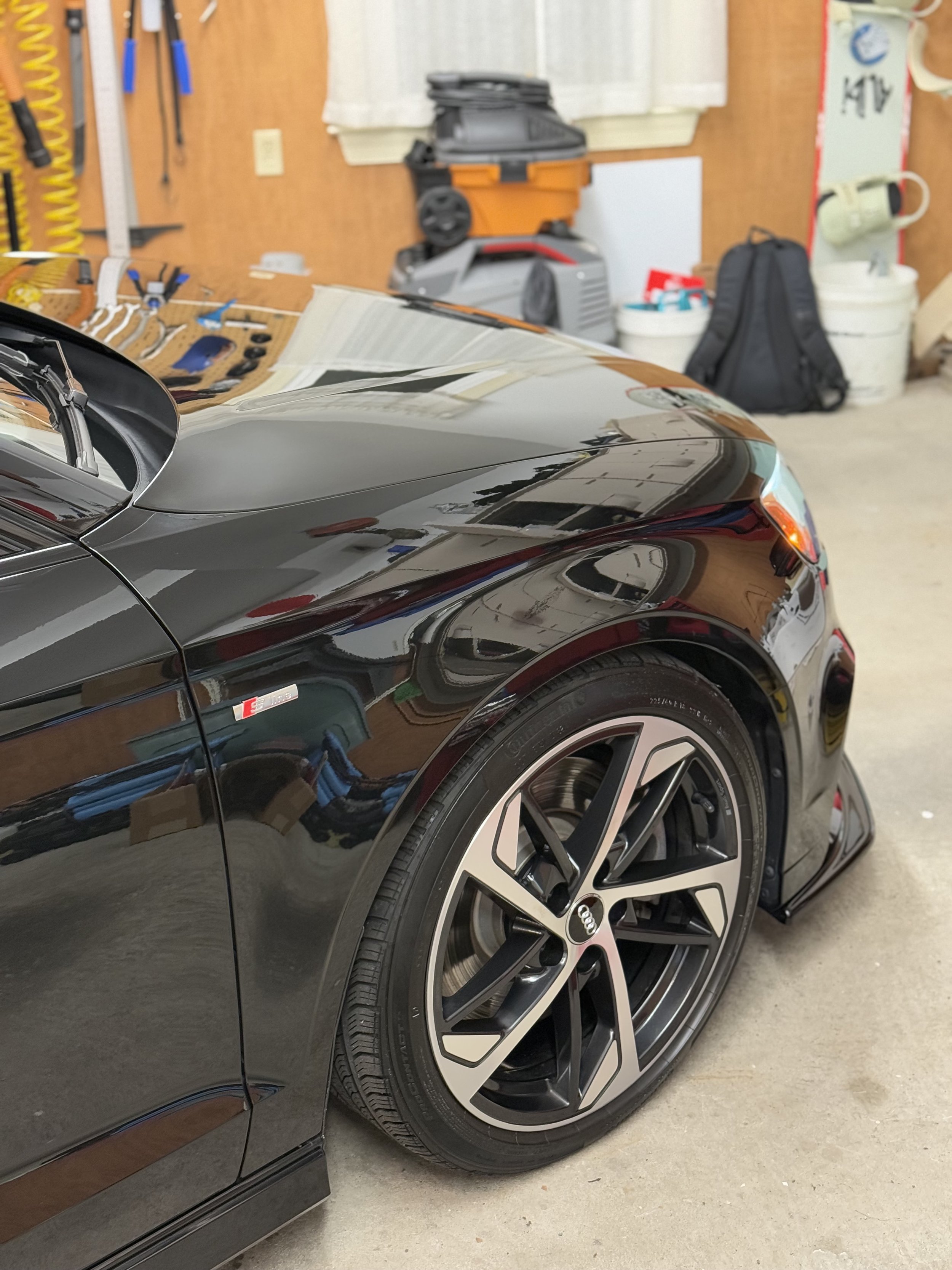 Close-up of a black Audi sports car's front right side with a modern alloy wheel, parked in a garage with various tools and equipment in the background.